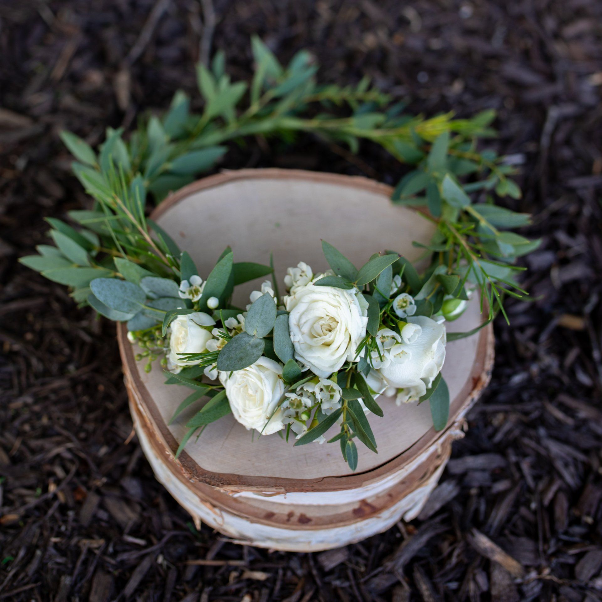 White floral wreath on stacked rustic bowls with green leaves, arranged on dark mulch