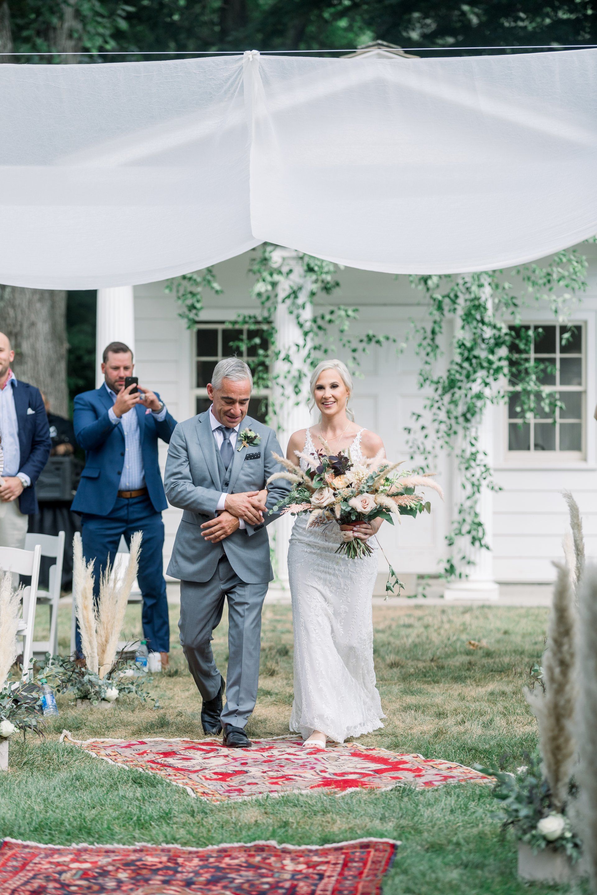 Bride and escort walking under a white canopy at an outdoor wedding ceremony, bouquet in hand