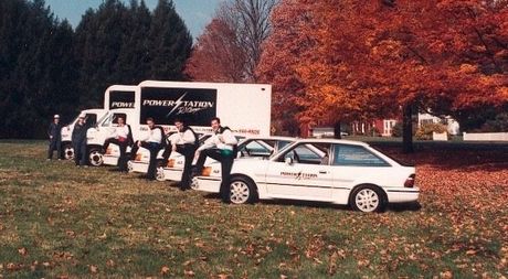 Workers unloading supplies from a white truck beside a white car in a grassy park in autumn