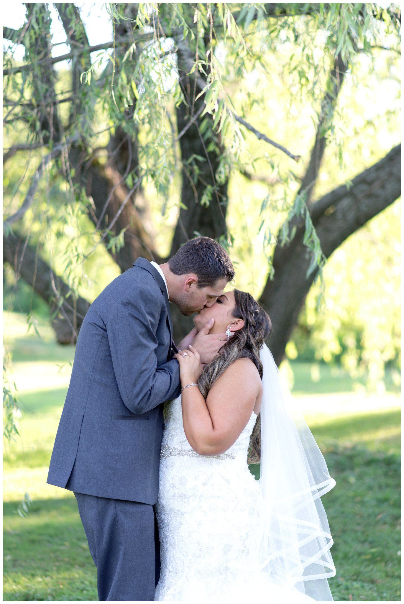 Couple in wedding attire kissing outdoors under trees, with a white veil visible.