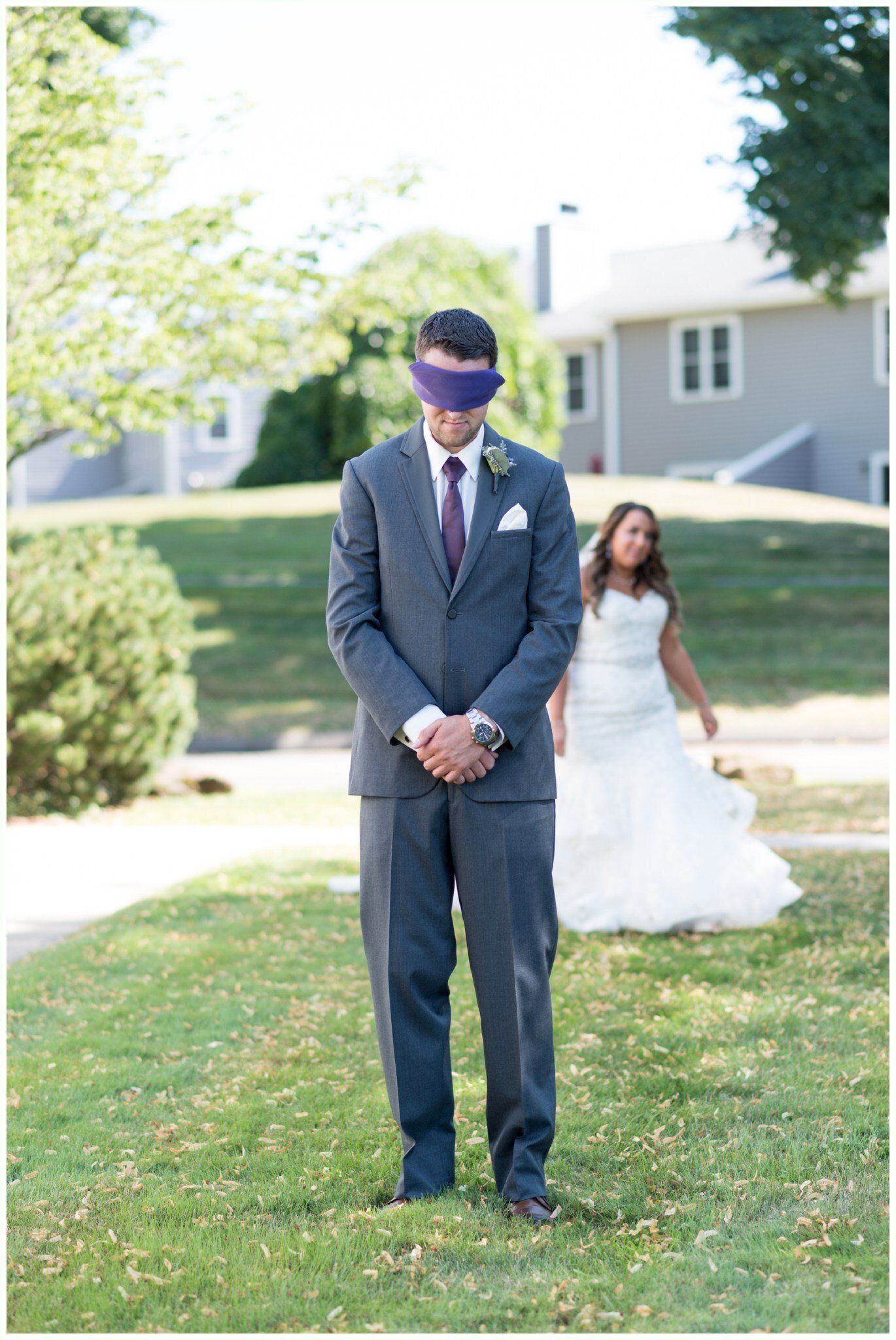 Man in a navy suit stands on grass with head bowed; bride in white dress smiles behind him outdoors.