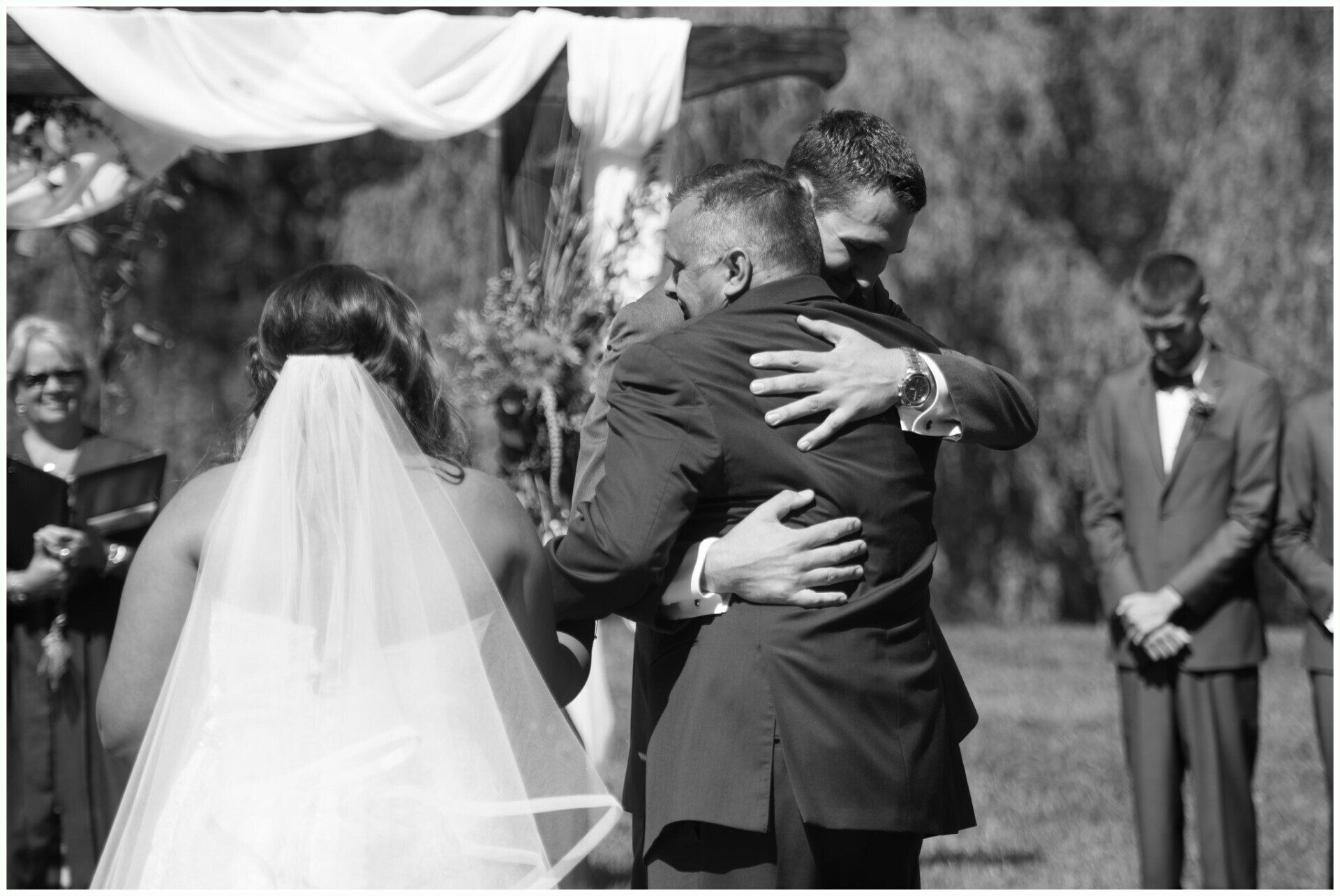 Bride in white veil embraces a man at an outdoor wedding ceremony, with guests watching nearby