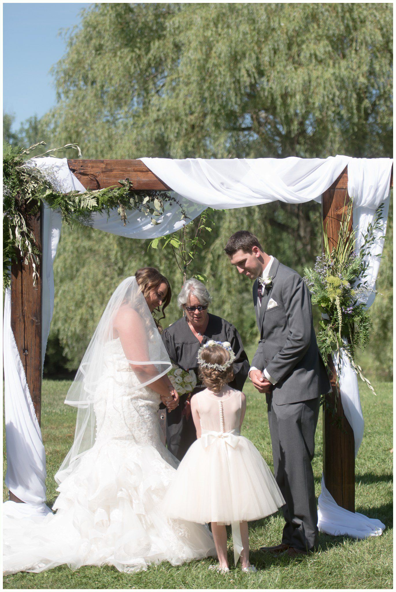 Wedding ceremony under a white-draped arch in a garden, with bride, groom, officiant, and child.