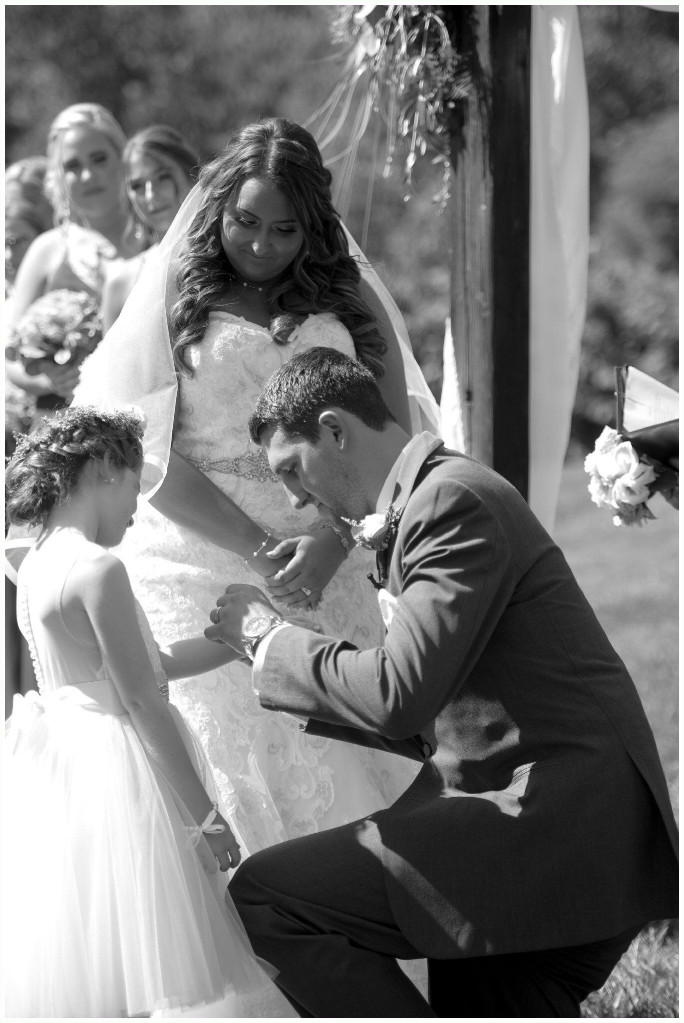 Man kneeling to place a ring on a bride's finger during an outdoor wedding ceremony