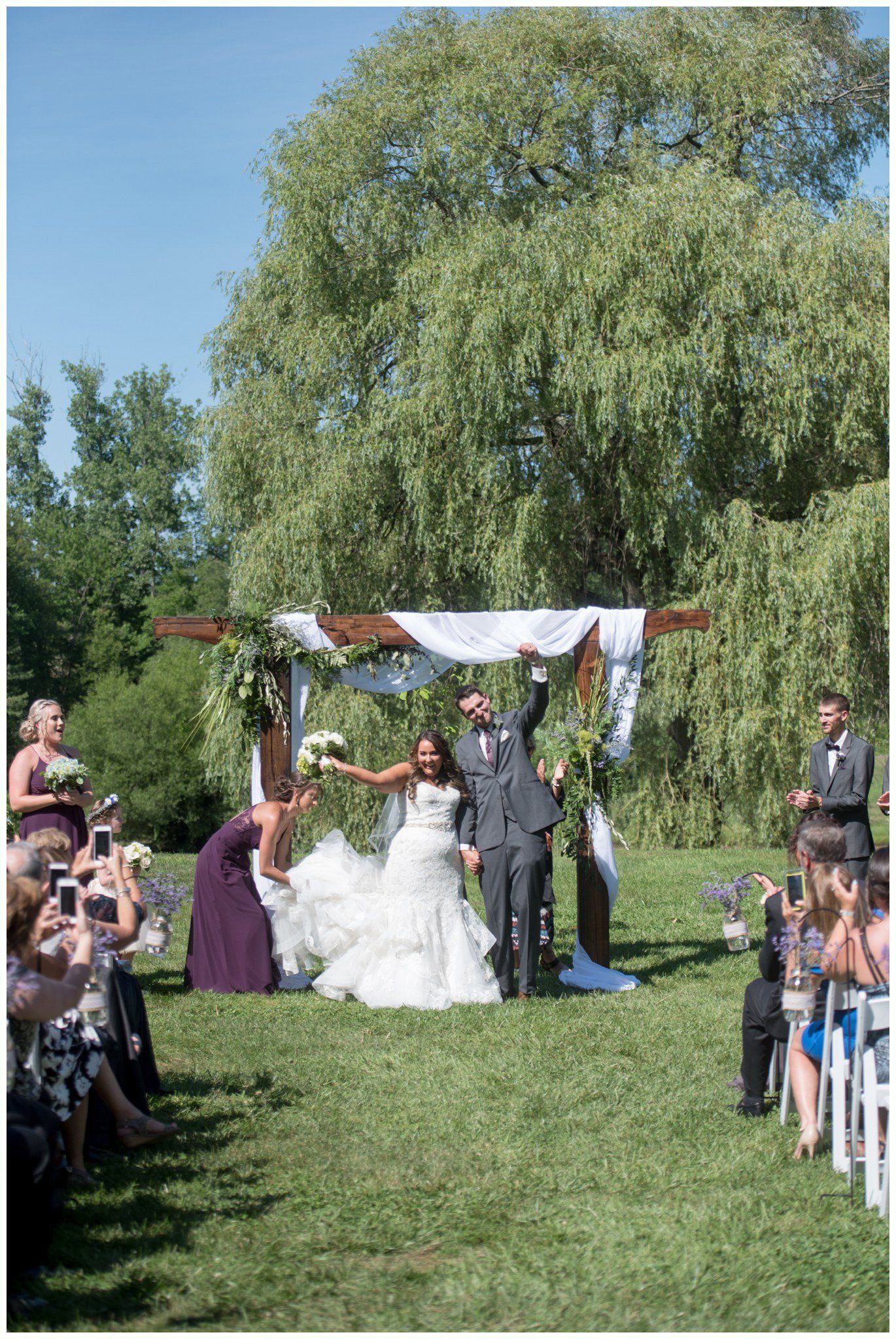 Wedding couple beneath a white arch on a grassy aisle, guests seated on both sides under a blue sky.