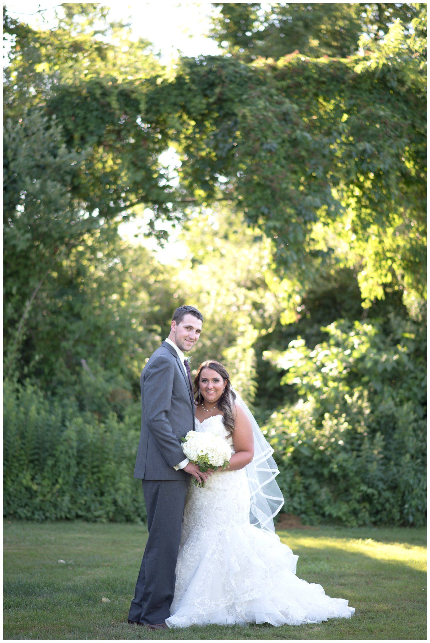 Bride and groom posing in a lush garden under trees, holding a bouquet.