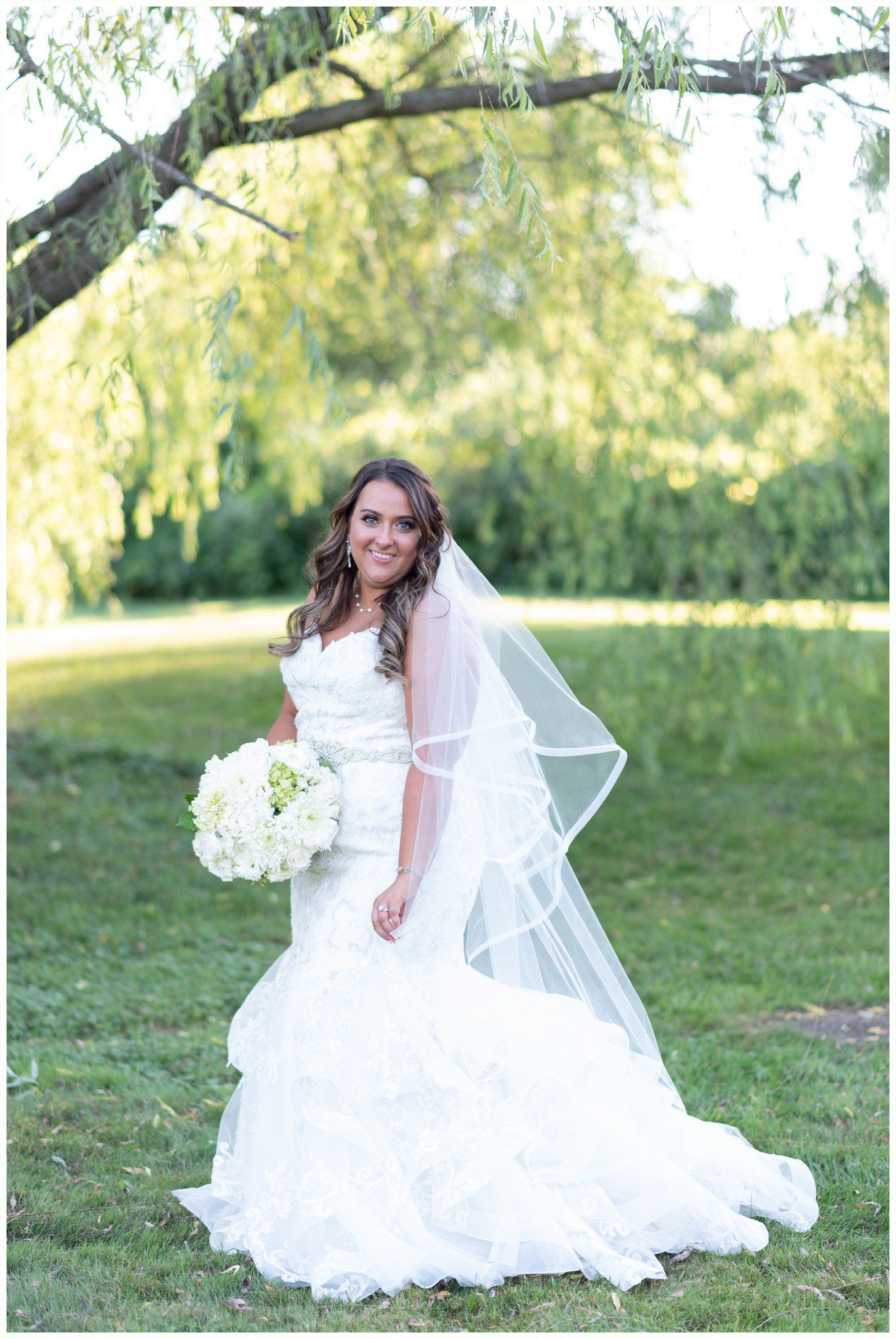 Bride in a white wedding dress standing on a grassy lawn holding a bouquet, with trees in the background