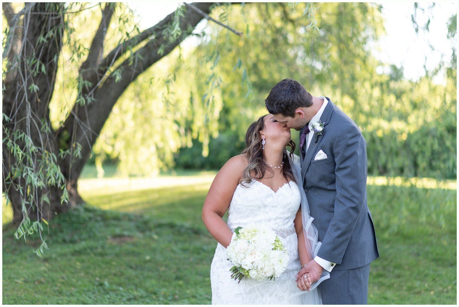 Bride and groom kiss in a sunlit park beside a large tree, bride holding a white bouquet.