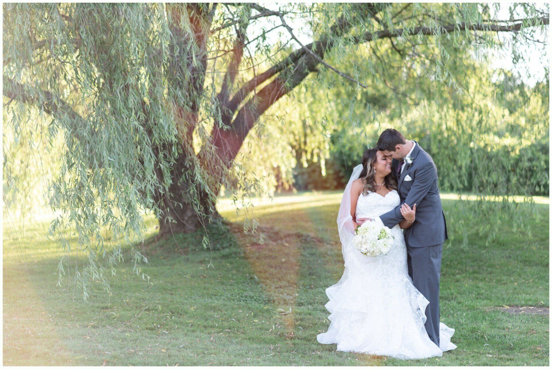 Bride and groom embracing under a willow tree in a sunlit park, with a bouquet of white flowers.