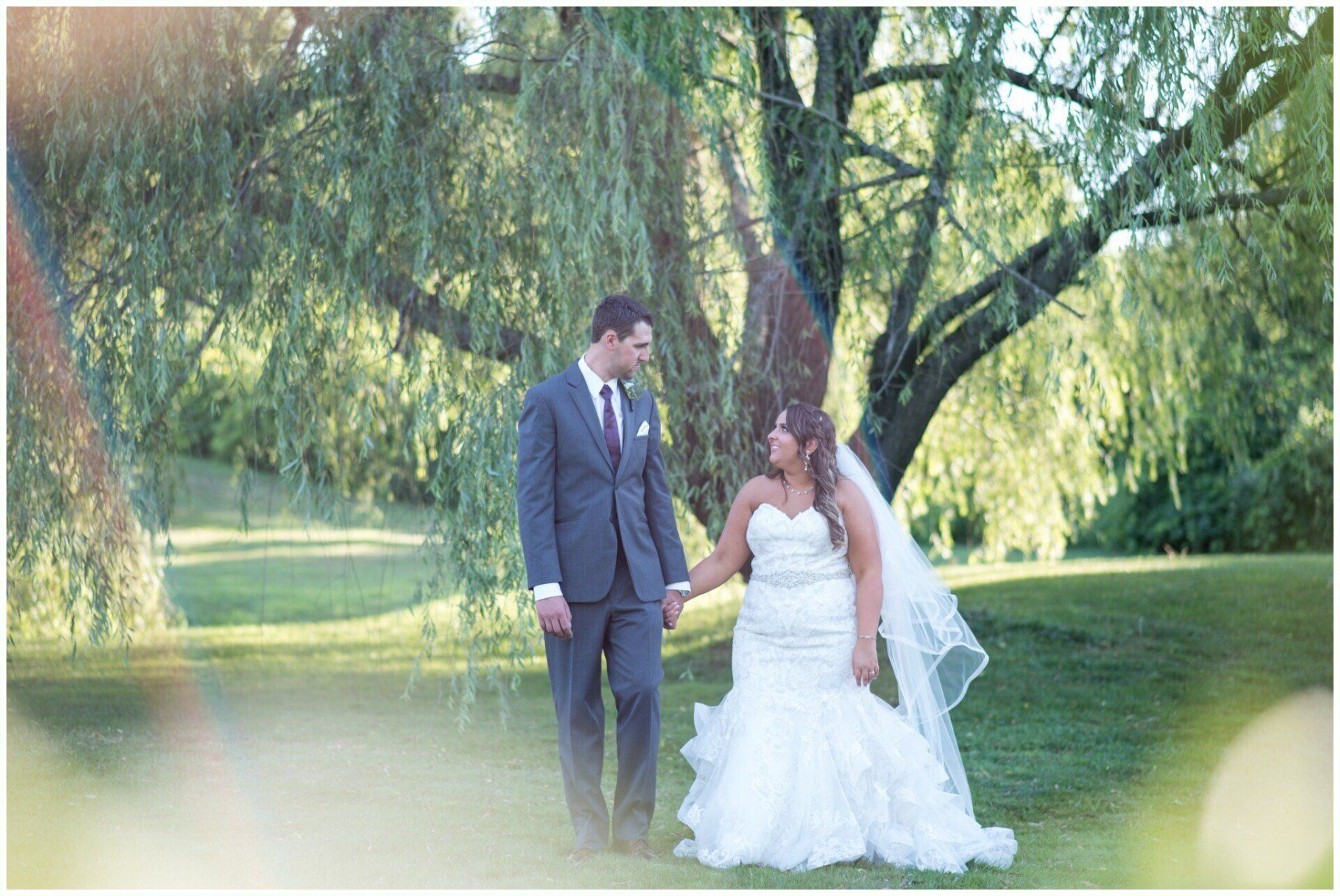 Bride and groom holding hands under a willow tree in a sunlit park