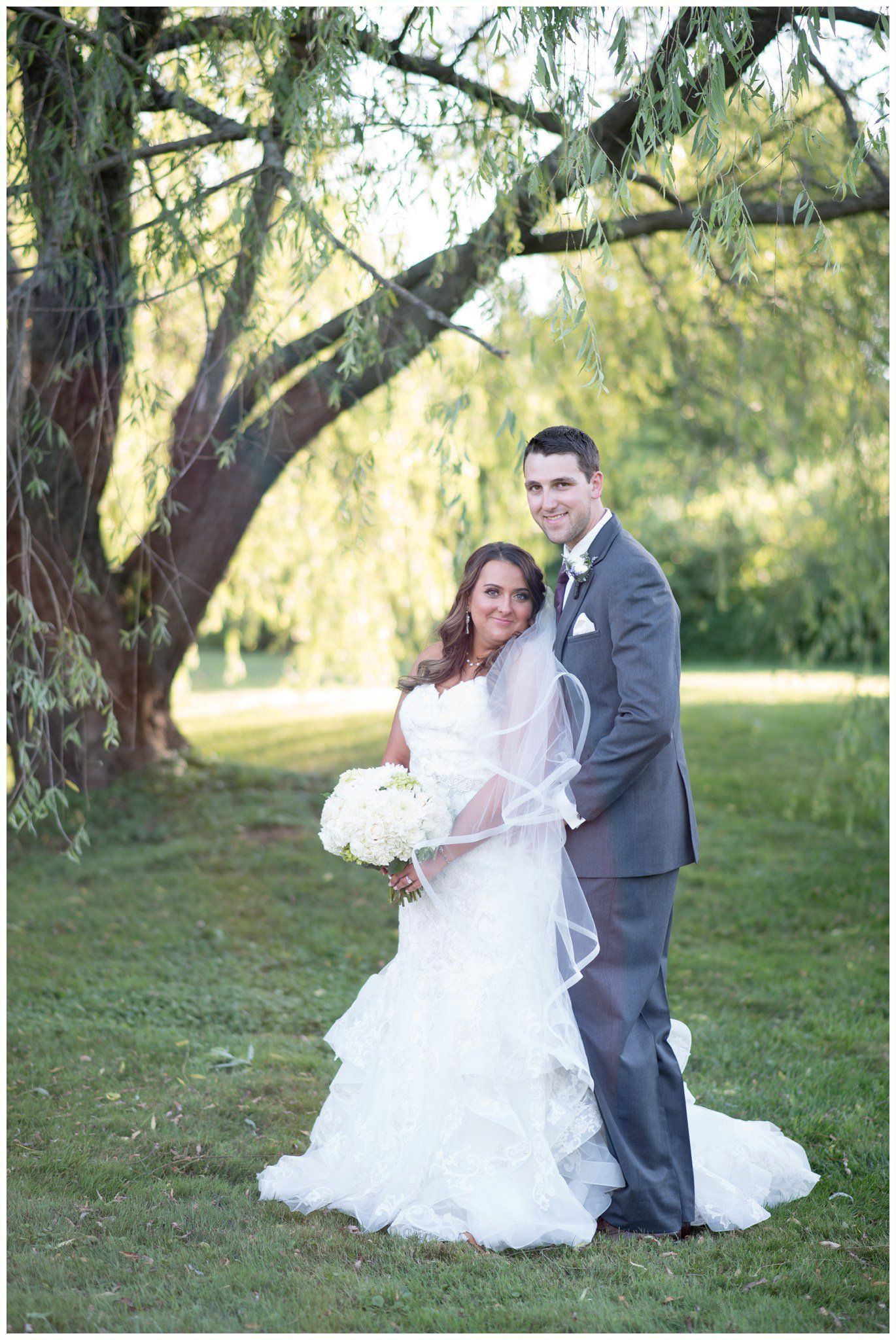 Bride and groom posing outdoors by a tree, holding each other on a grassy lawn.