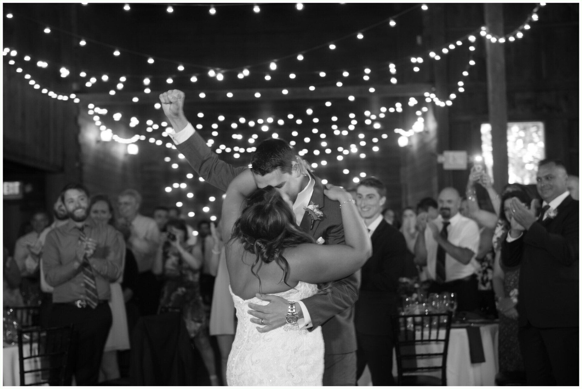 Newlyweds kiss under string lights while guests applaud at an outdoor evening reception