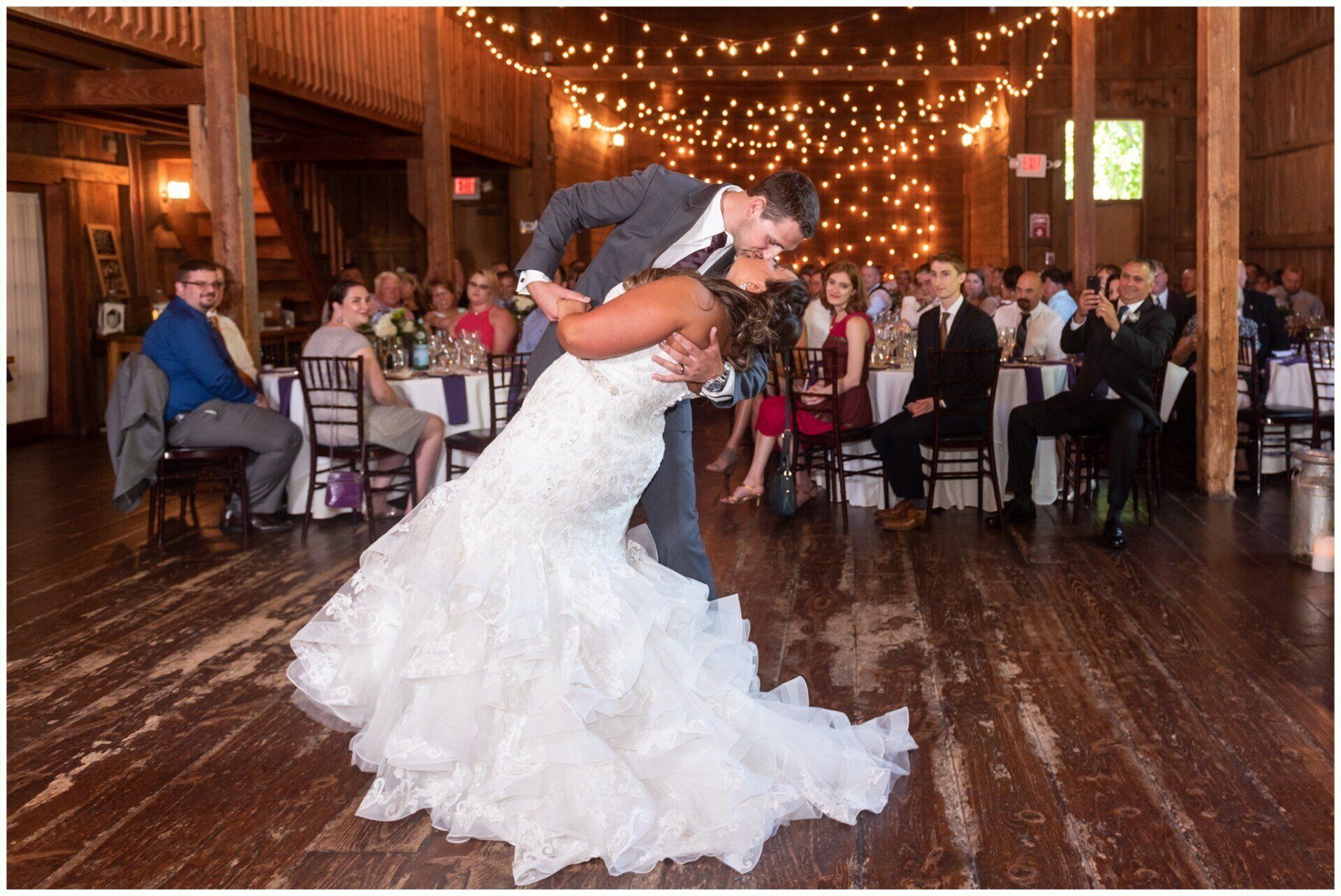 Bride and groom dip kiss on a wooden dance floor under warm string lights at a reception