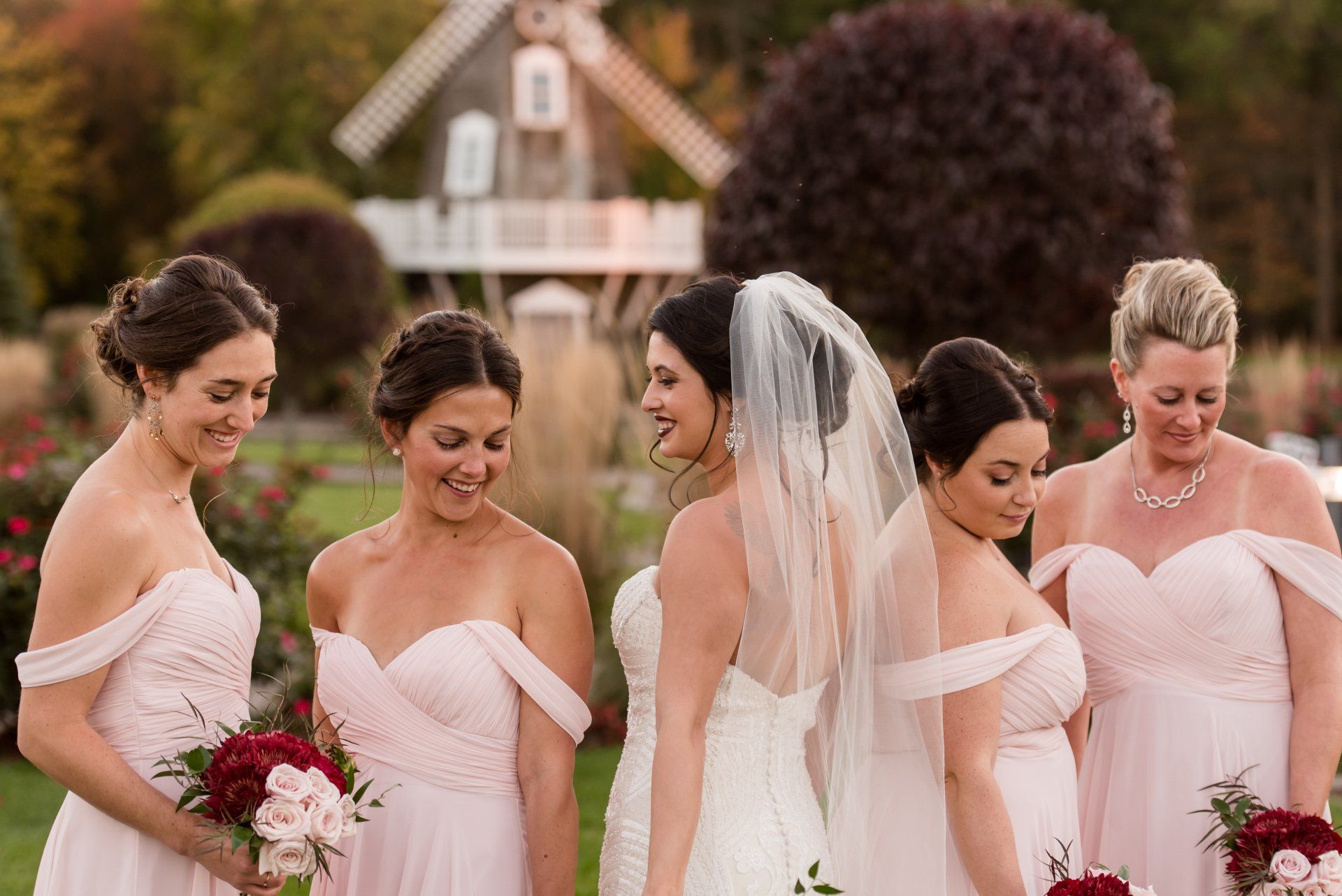Five bridesmaids in blush dresses and a bride in a veil stand outdoors holding bouquets.