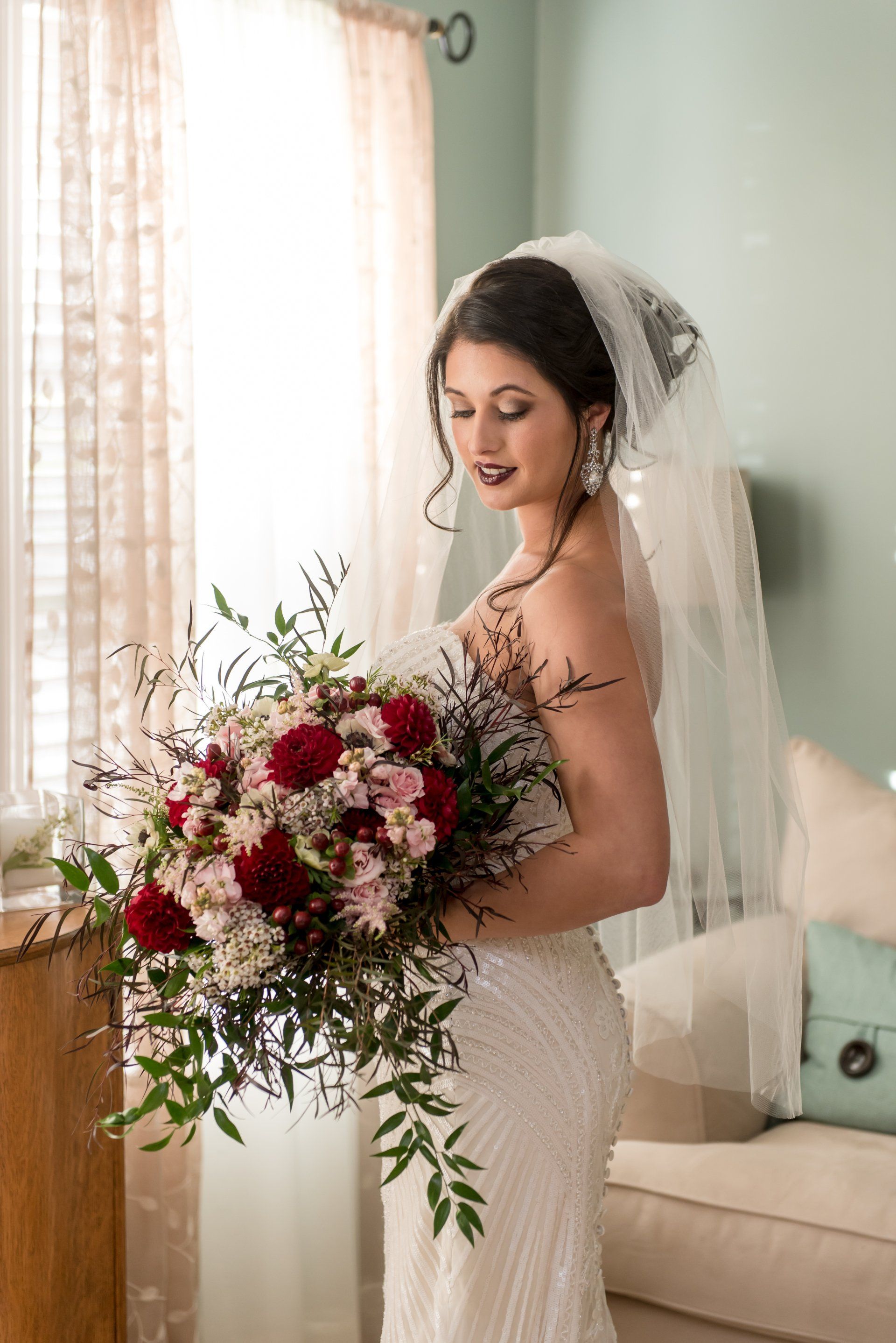 Bride in a lace gown holding a red and white bouquet beside a sunlit window