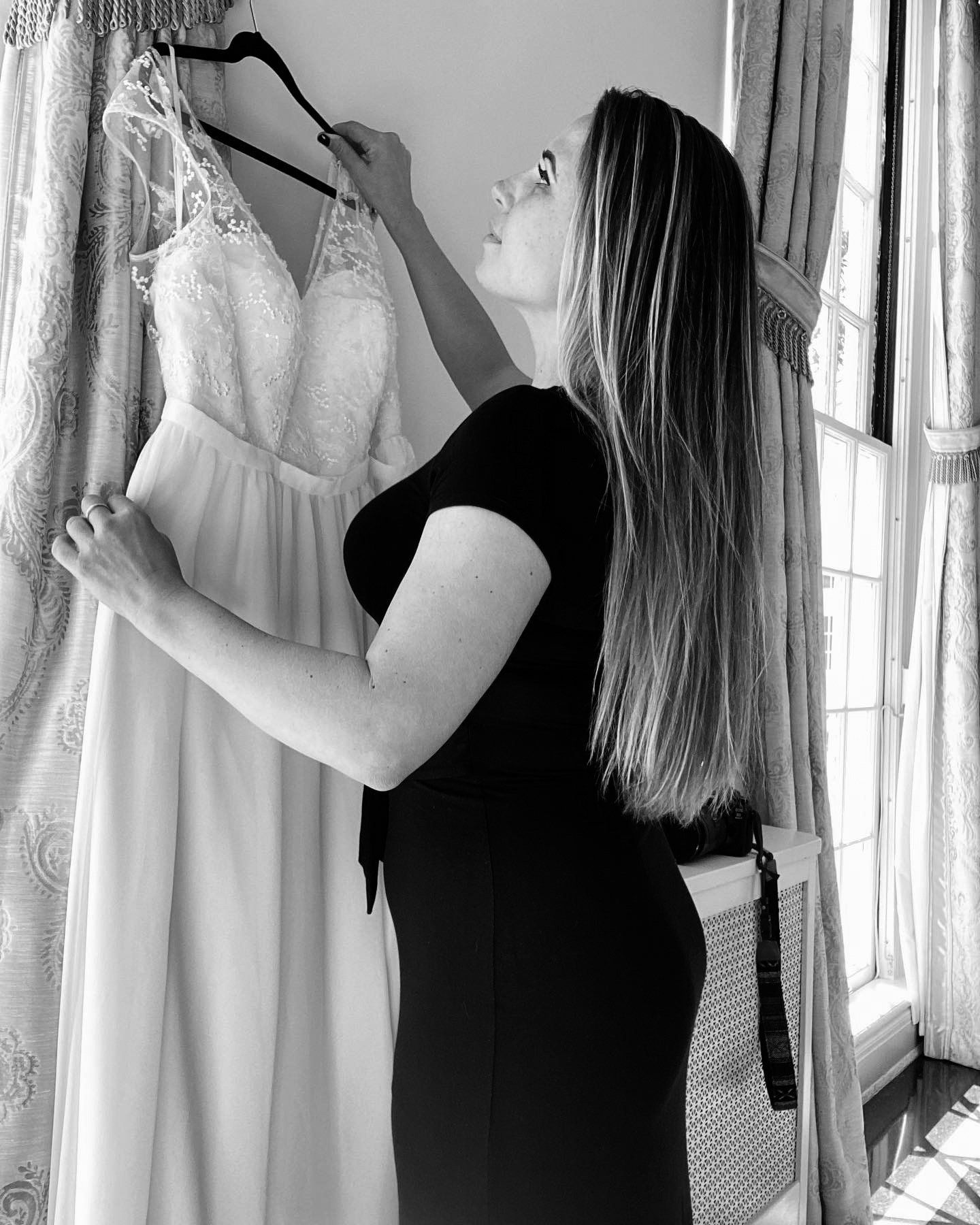 Woman in a black dress adjusting a white lace gown on a hanger by a window