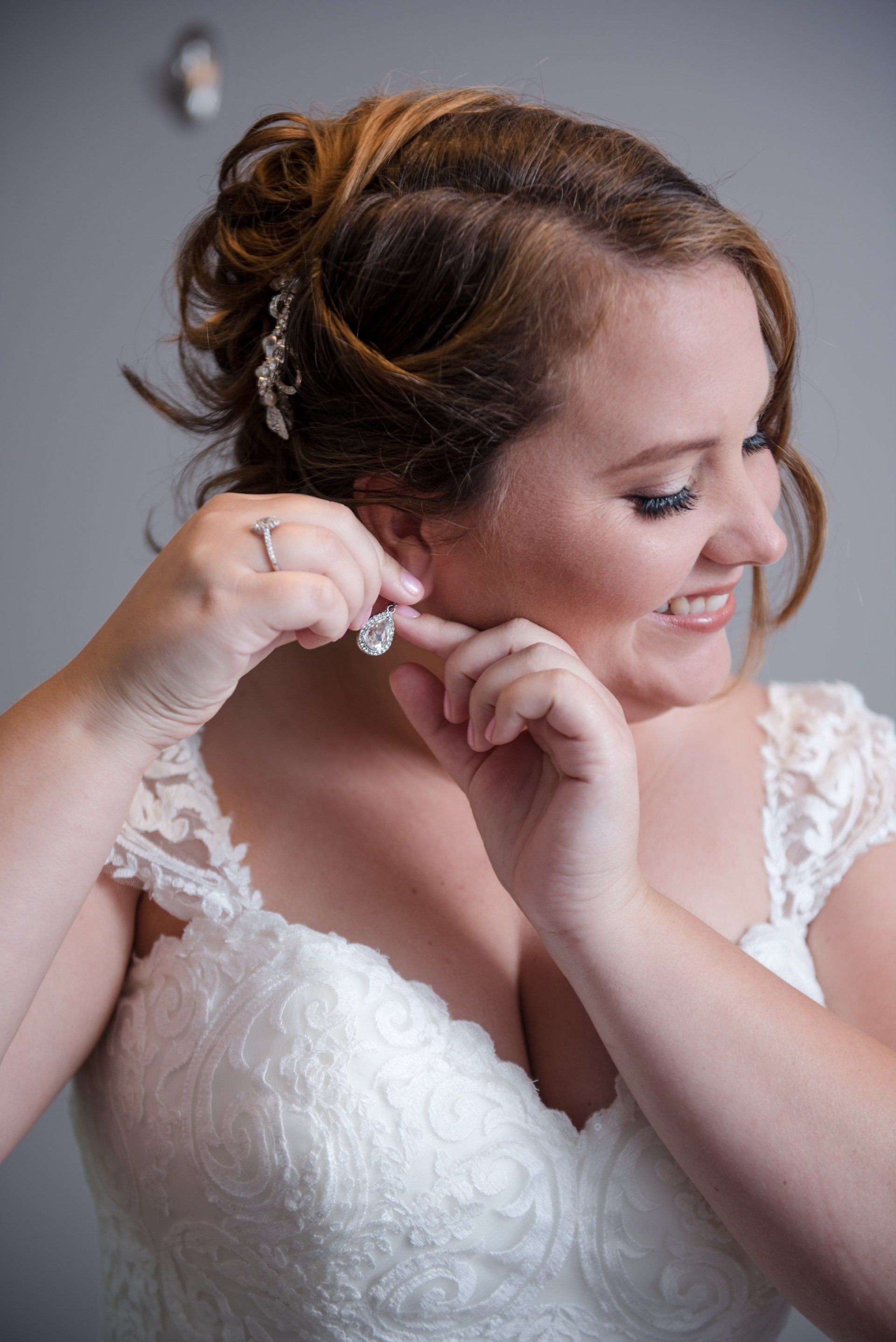 Bride fastening an earring, smiling in a white lace wedding dress