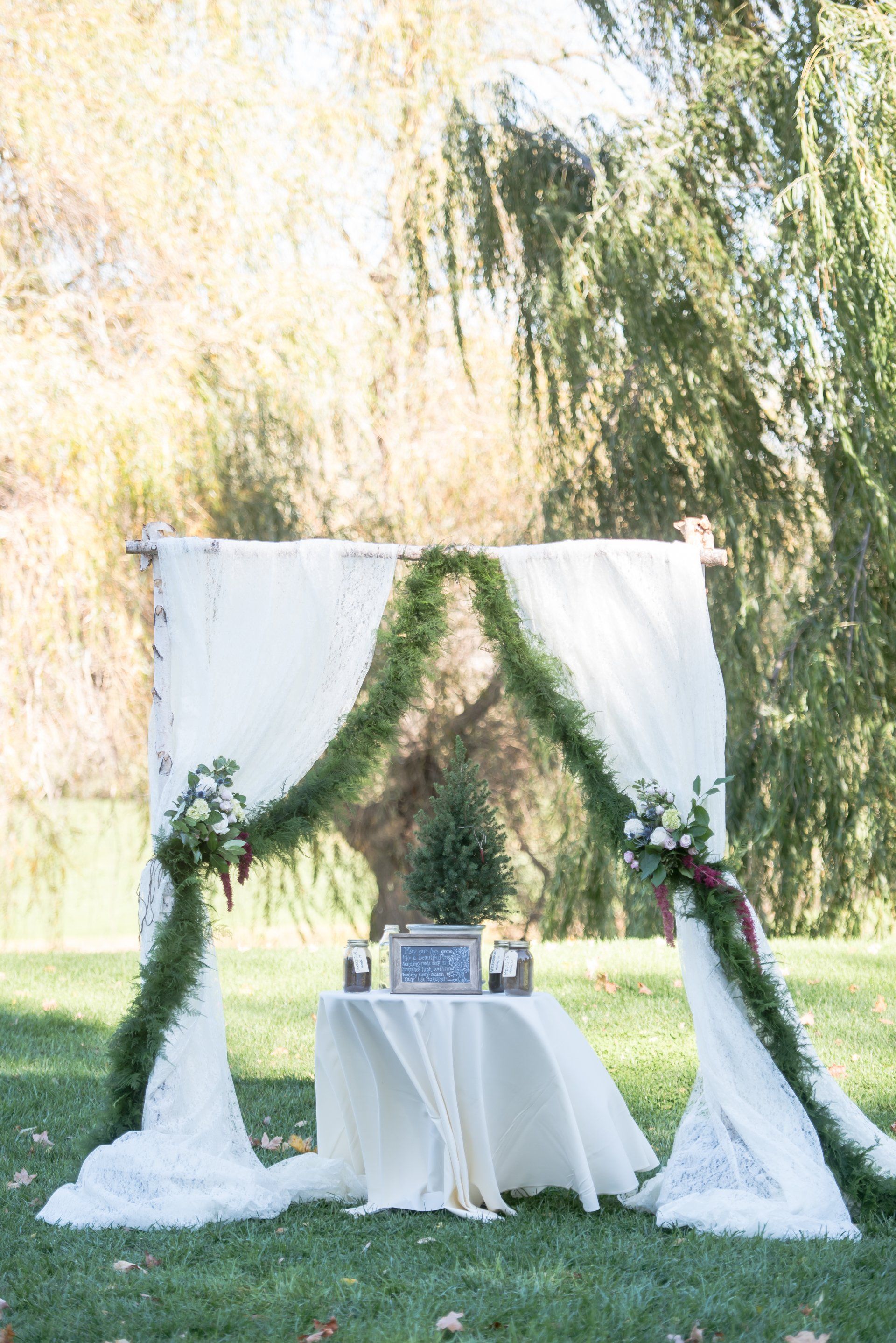 White draped wedding arch with greenery and a small table in a sunlit garden
