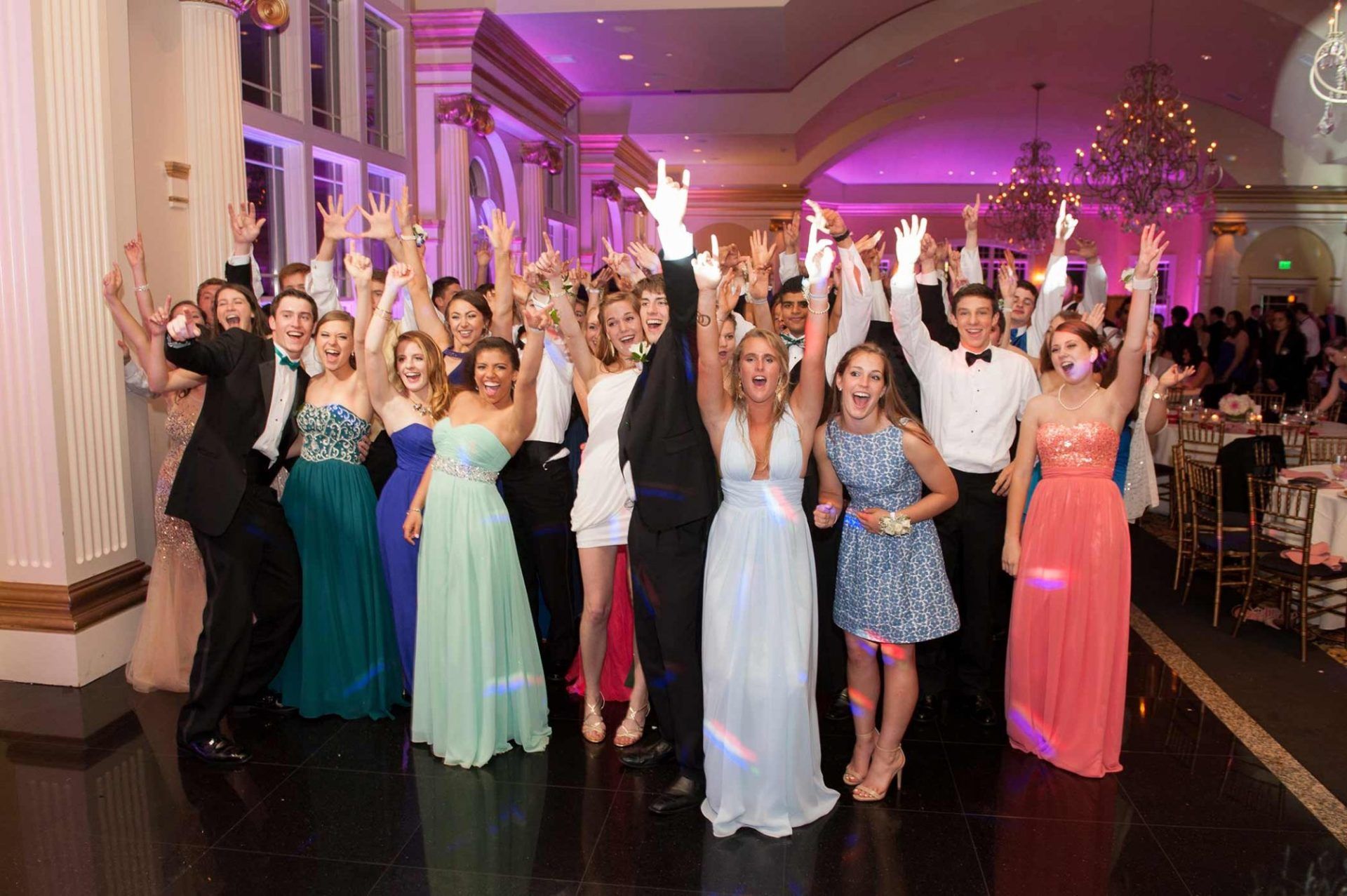 Group of people in formal dresses and suits cheering with raised arms at a ballroom event.