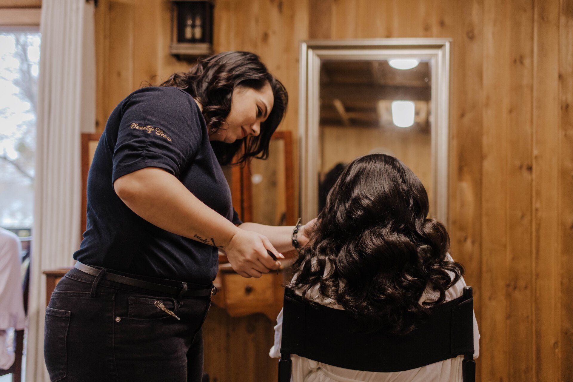 Stylist curling a seated person’s hair in a wood-paneled room with mirror and warm lighting