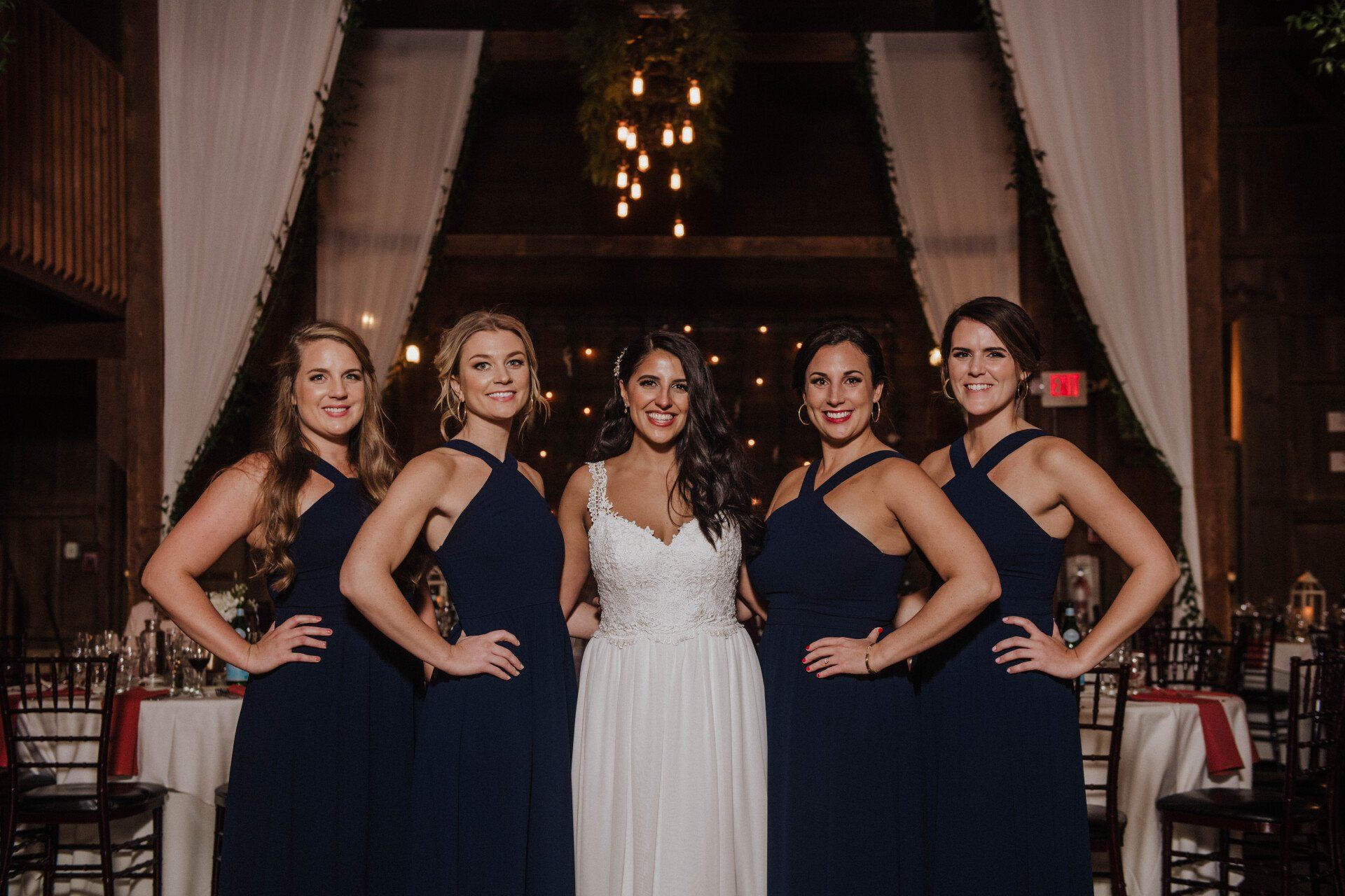 Five women in formal dresses pose in a decorated hall, with one in white centered between four in navy.