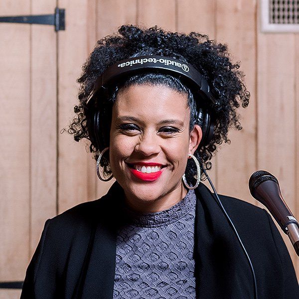 Smiling woman wearing headphones stands beside a microphone against a wooden wall.