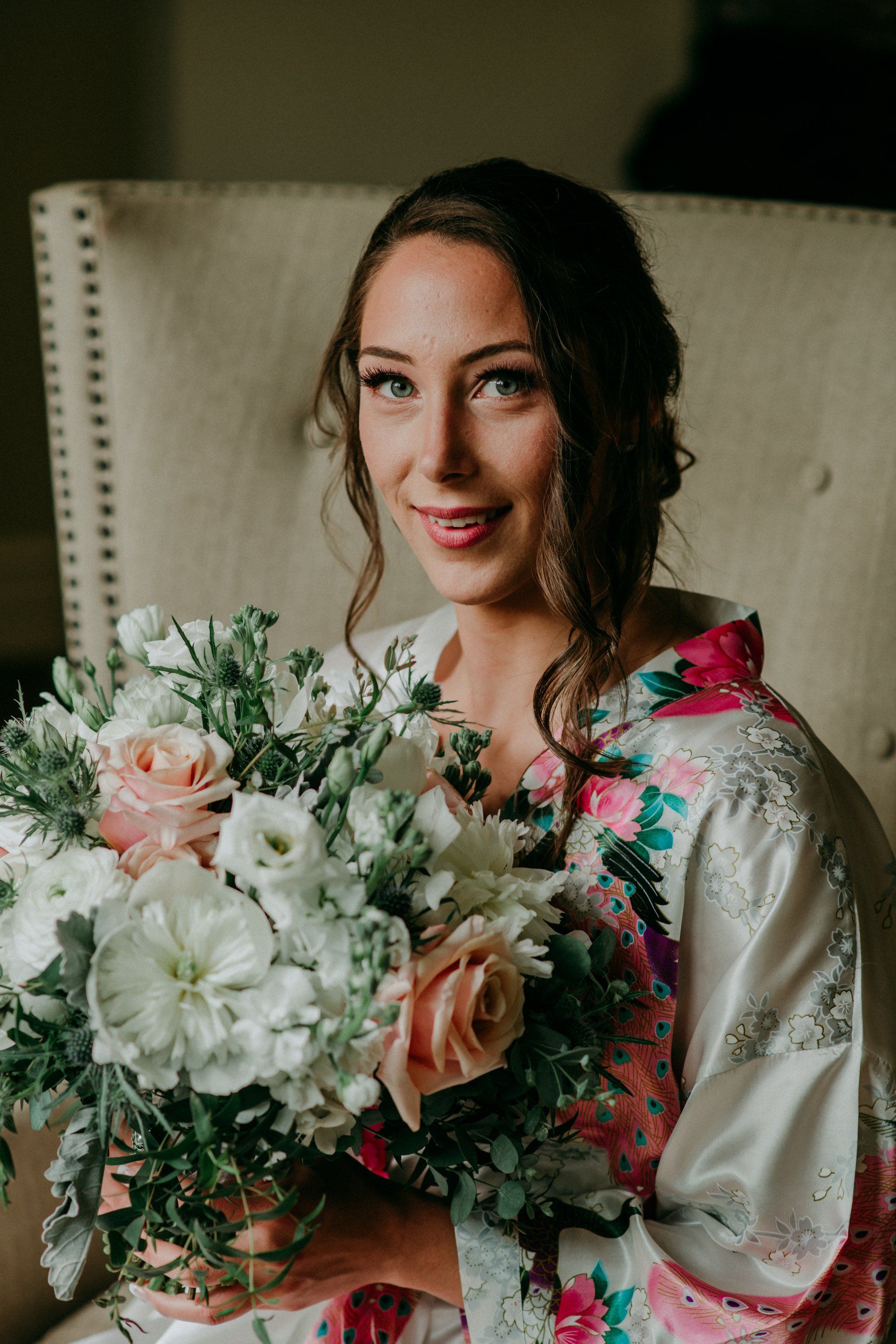 Woman in a satin robe holding a large bouquet of white and pink flowers, seated in a cream chair.