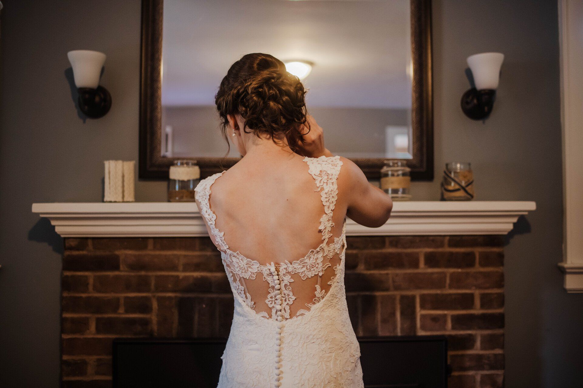 Bride in a white lace-back dress adjusting her reflection at a mantel mirror in a dim room