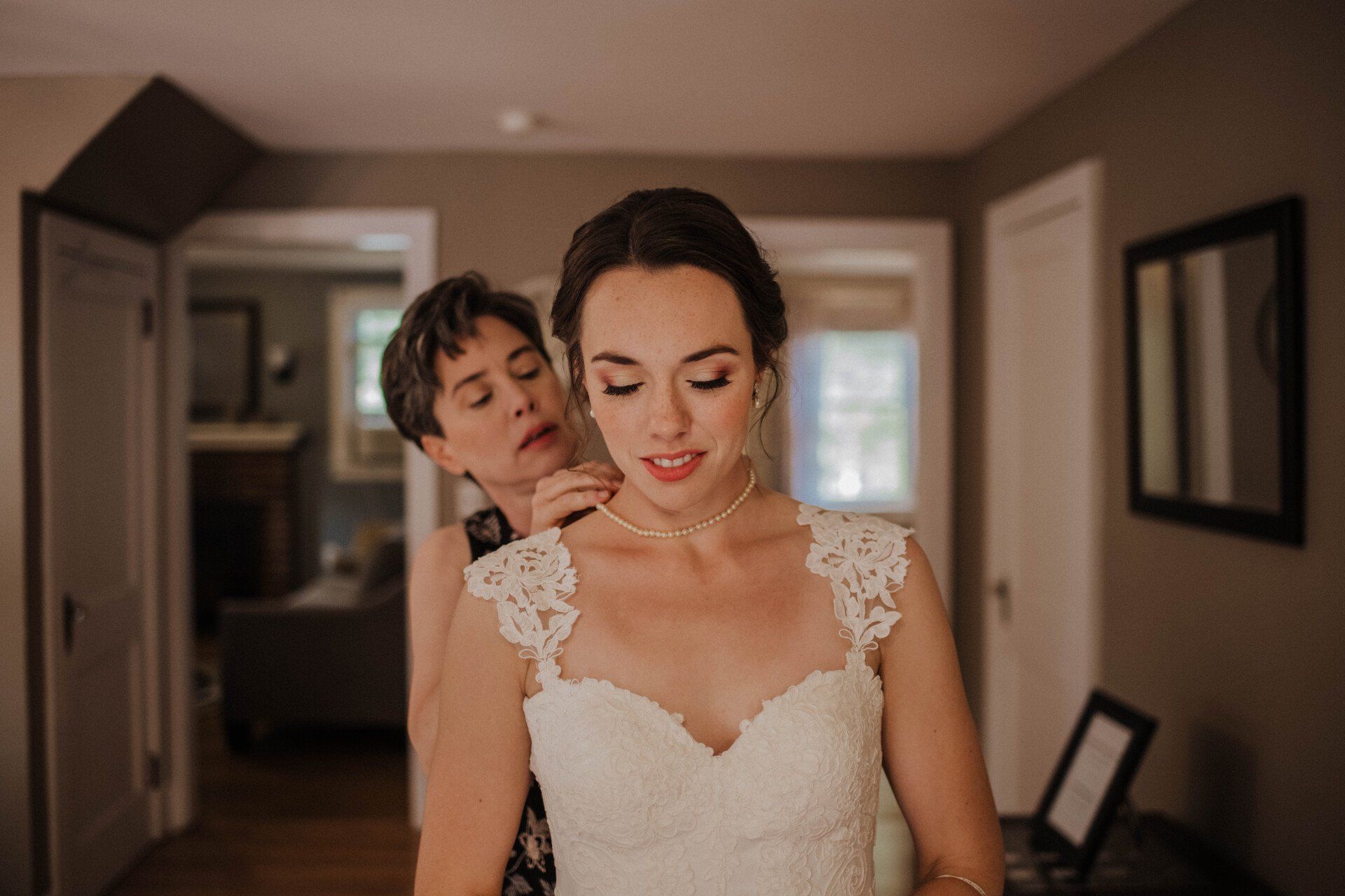 Bride adjusting necklace in doorway, with a helper behind her in a softly lit hallway.