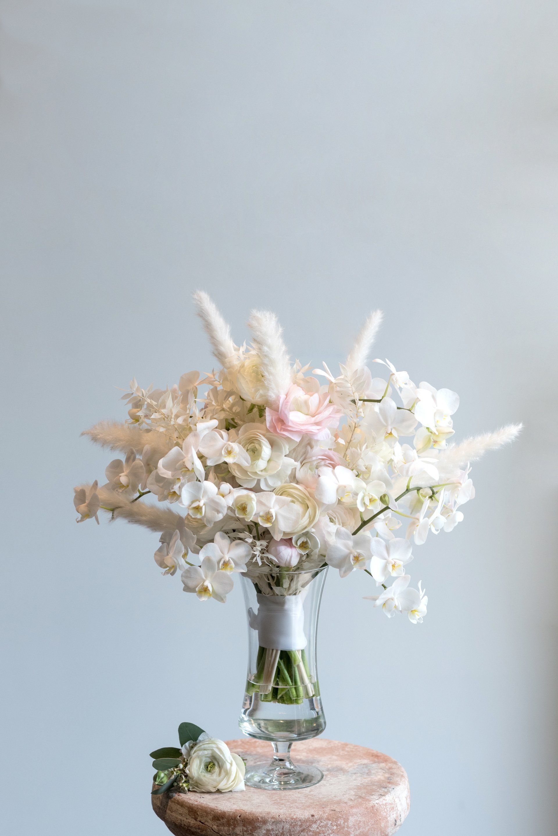 A small glass vase of pale white and pink flowers on a pedestal against a light gray background