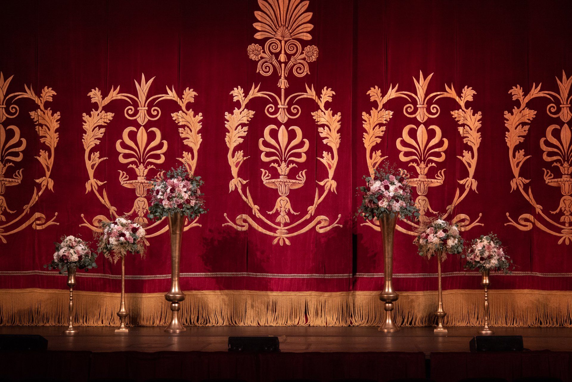 Red theater stage with ornate crimson curtain and gold trim, flanked by floral arrangements and microphones