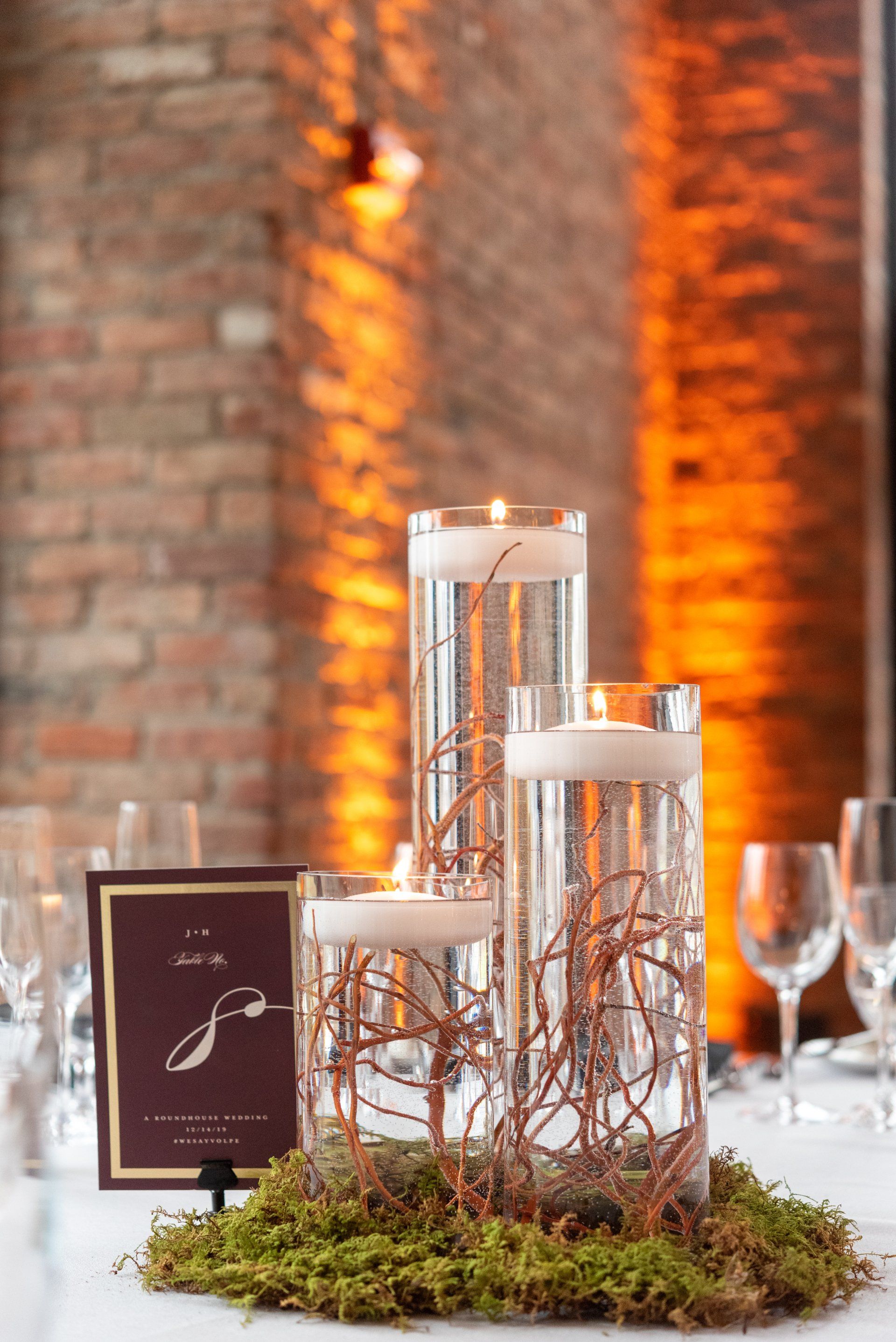 Three glass cylinder candles on moss with amber lights and a menu card on a table.