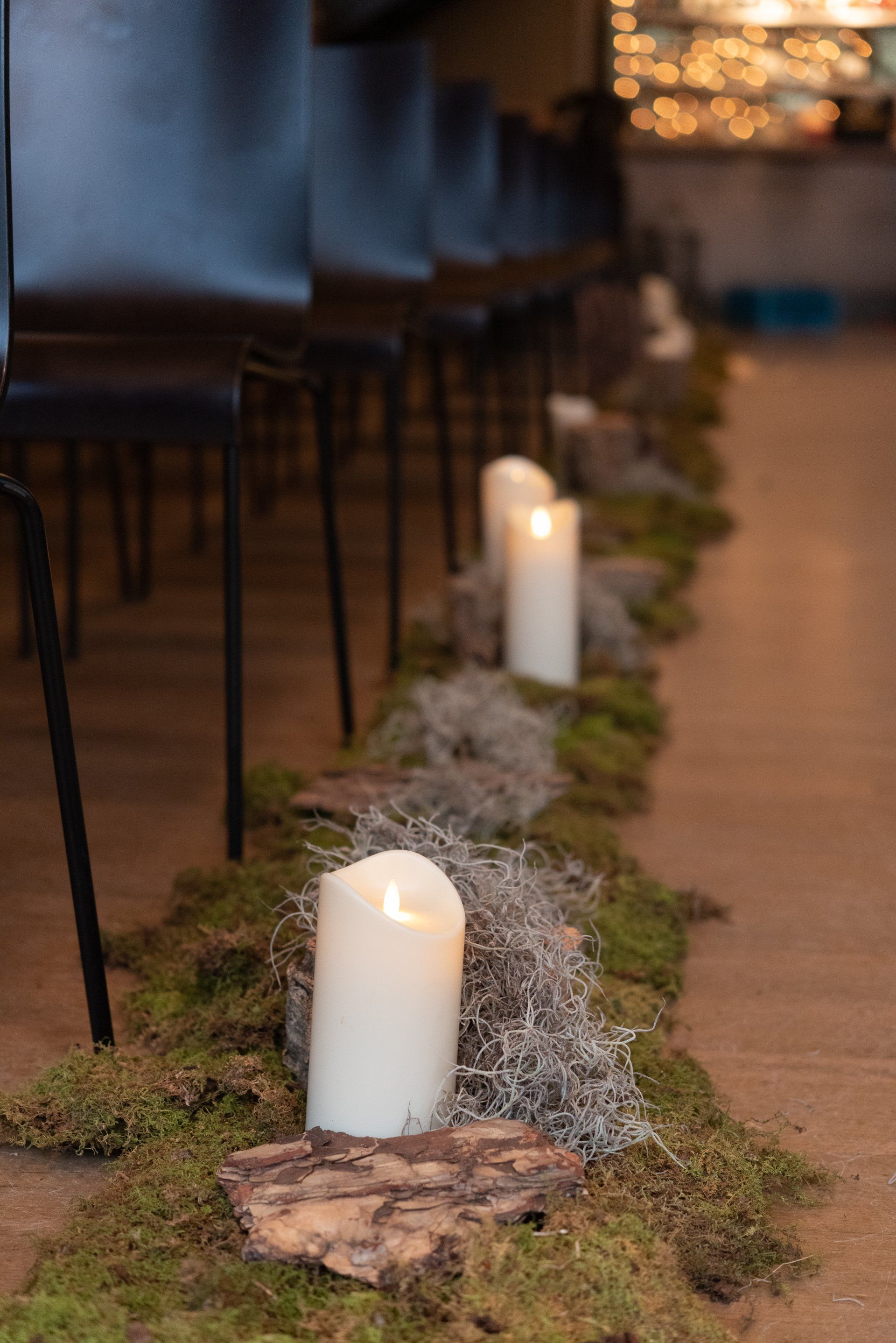 Candlelit aisle with white pillar candles and moss along the floor beside dark chairs