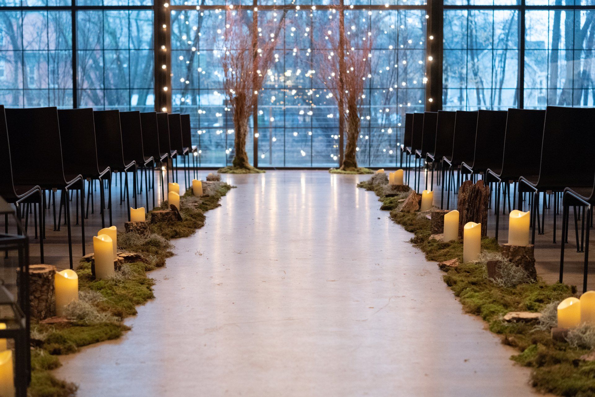 Indoor wedding aisle with candles, greenery, and black chairs facing tall windows and a floral arch.