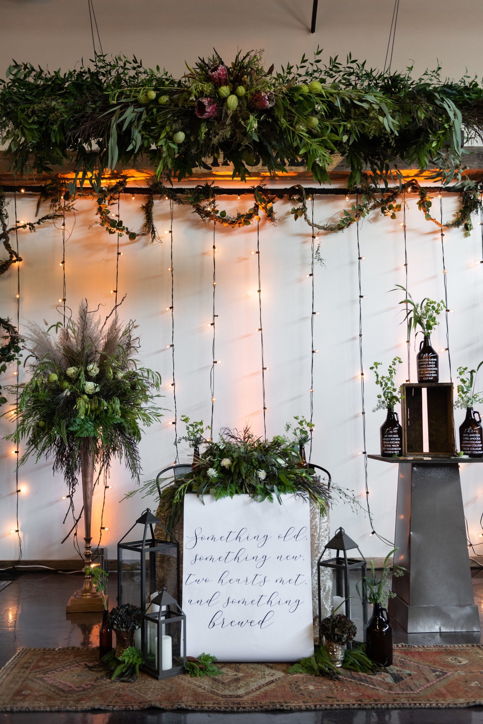 Decorative white wall with hanging greenery, fairy lights, lanterns, and a framed sign at a wedding-style display