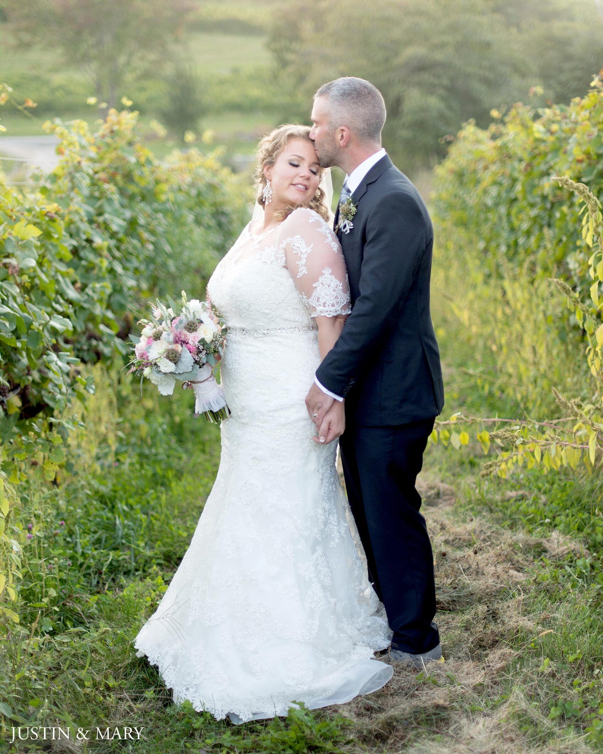 Bride in a white gown and groom in a dark suit embracing in a vineyard path at sunset