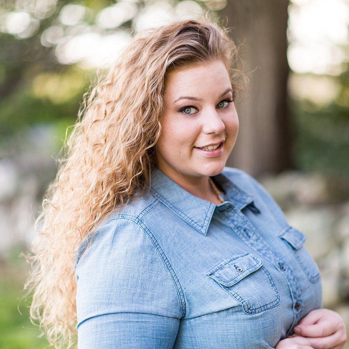 Smiling woman in a light blue denim shirt outdoors with a blurred natural background