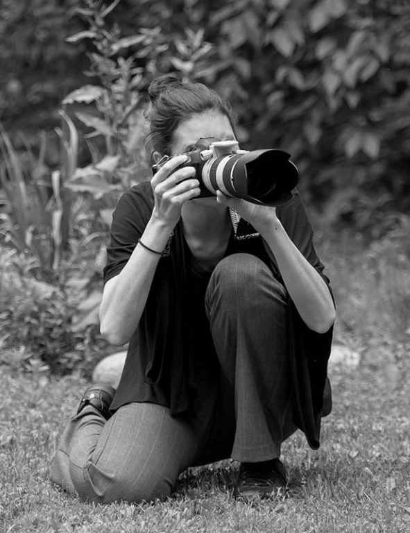 Person kneeling outdoors, aiming a camera with a large telephoto lens through plants