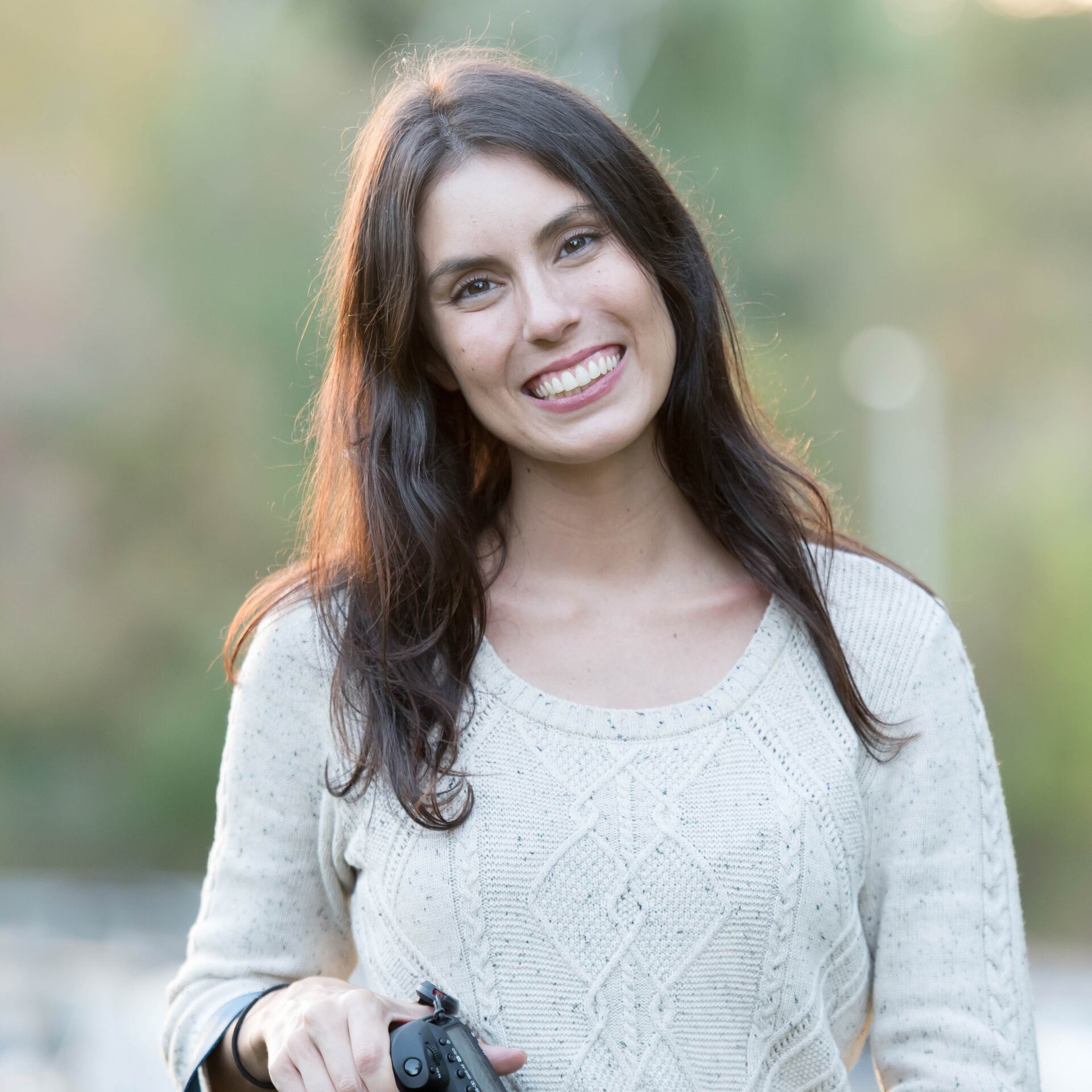 Smiling woman in a cream sweater holding a camera outdoors with blurred greenery behind her