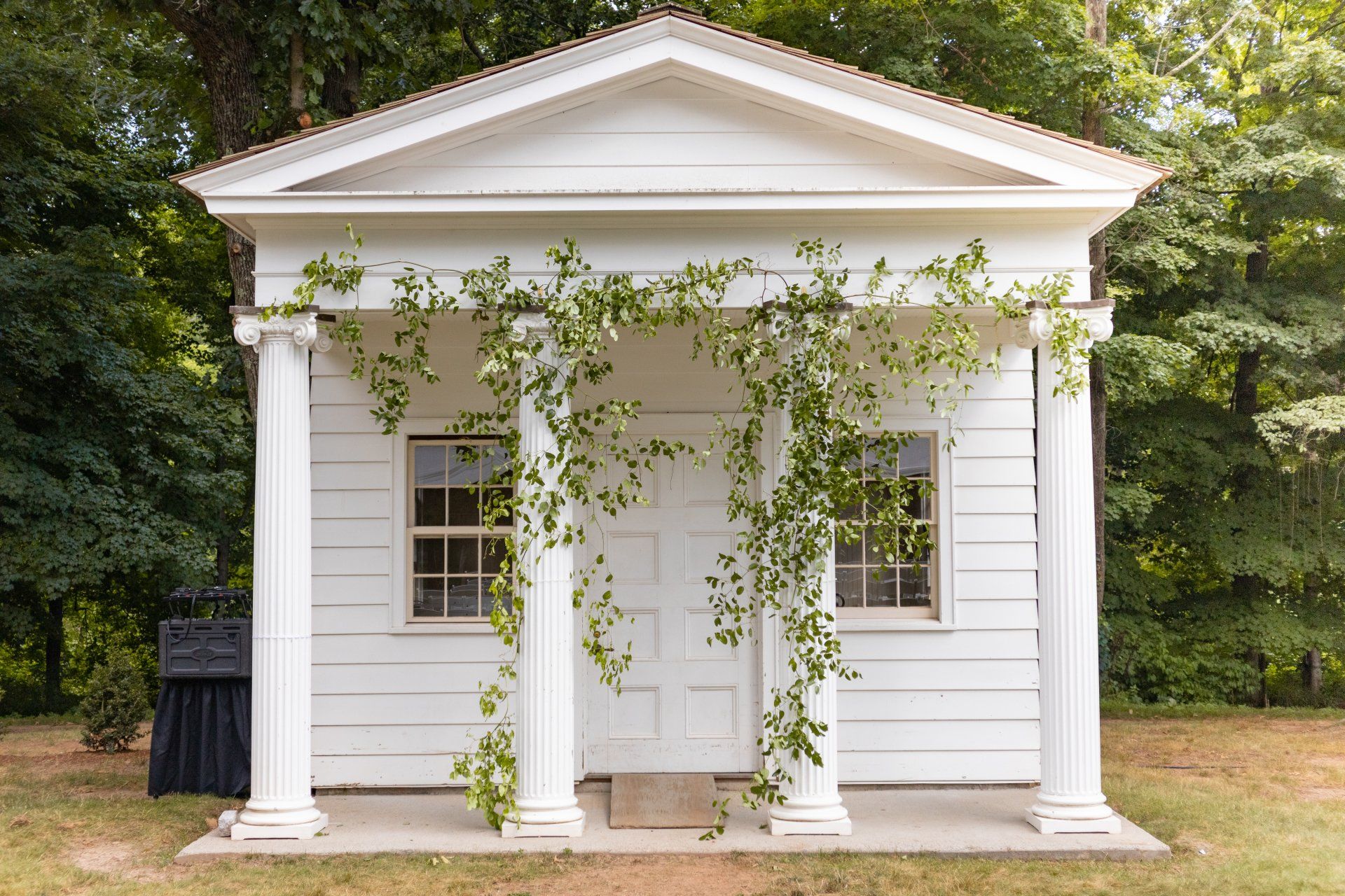 Small white neoclassical building with columns and vines at the entrance in a wooded setting