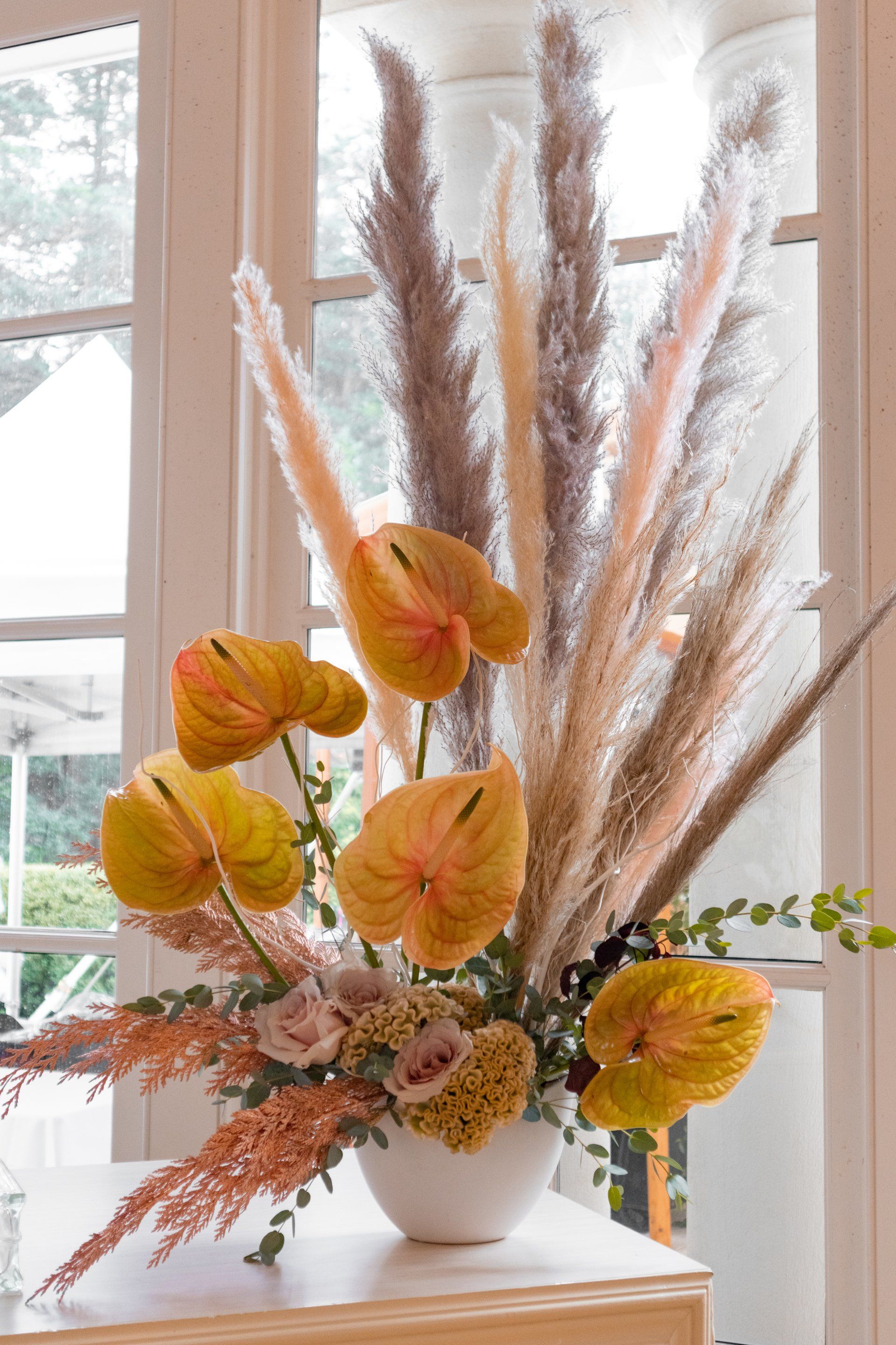 White vase of autumn floral arrangement with yellow flowers and tall beige pampas grass by a window