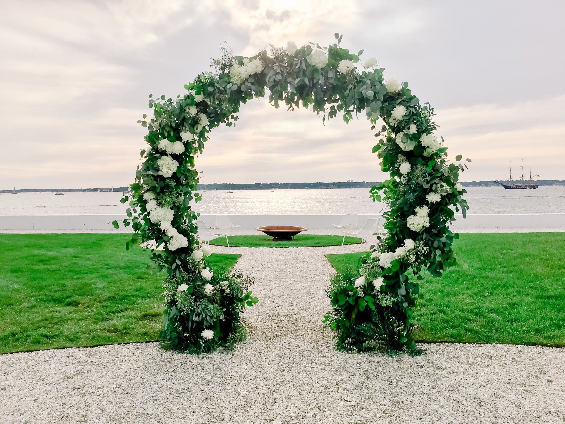 Floral arch on a gravel path by a grassy waterfront at sunset