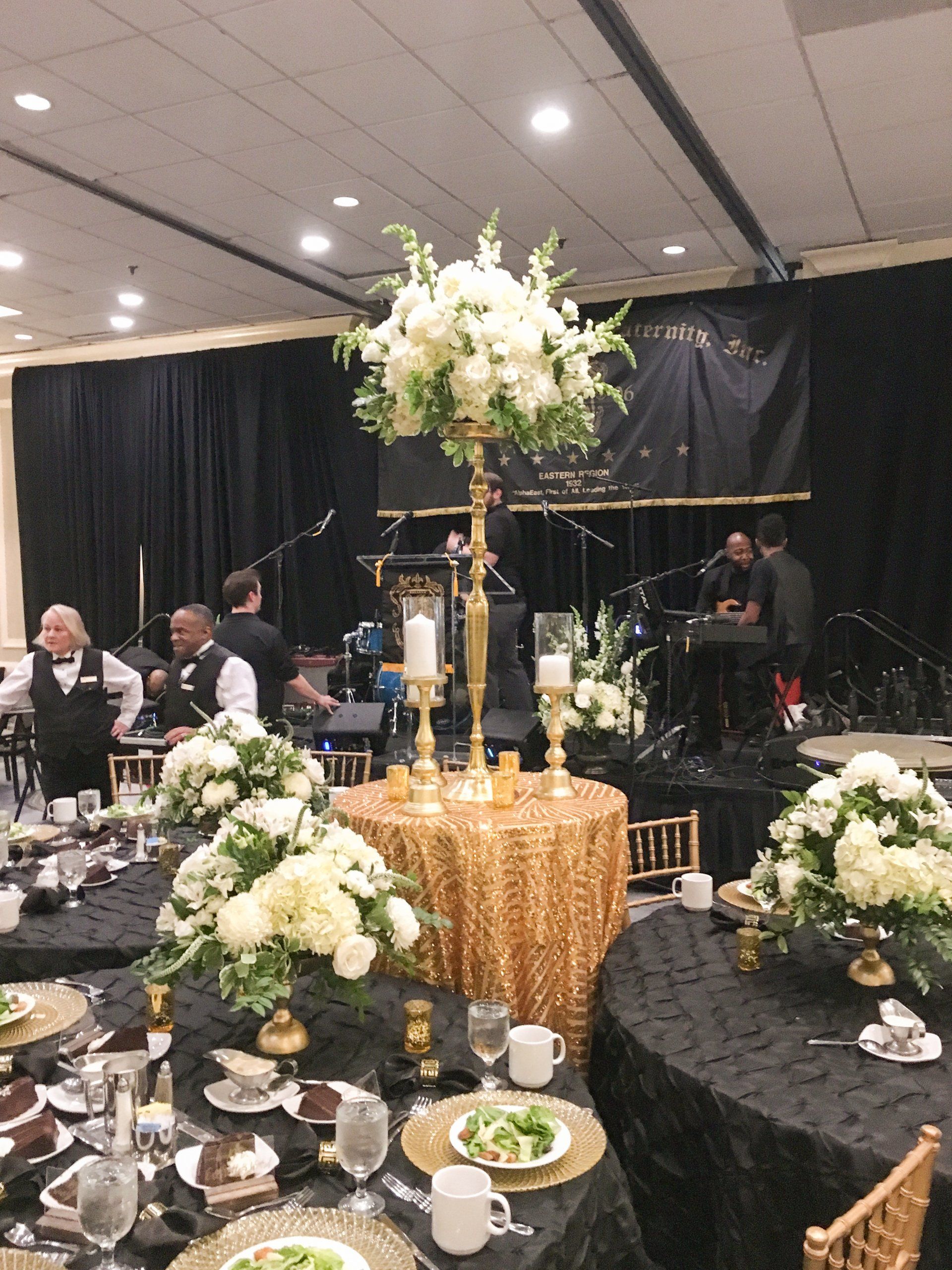 Decorated banquet hall with round tables, black-and-gold decor, and a tall white floral centerpiece near a stage