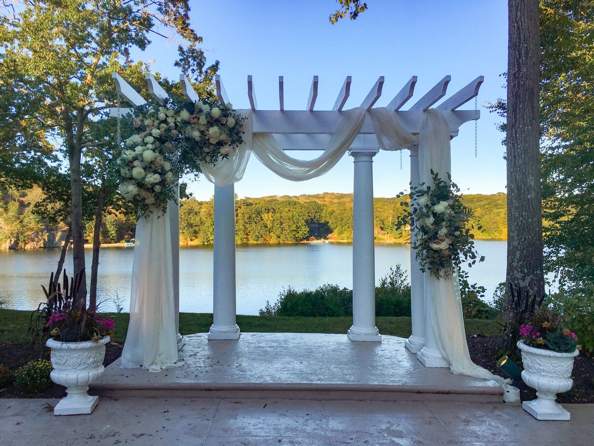 White wedding arch with draped fabric and flowers on a lakeside patio at sunset