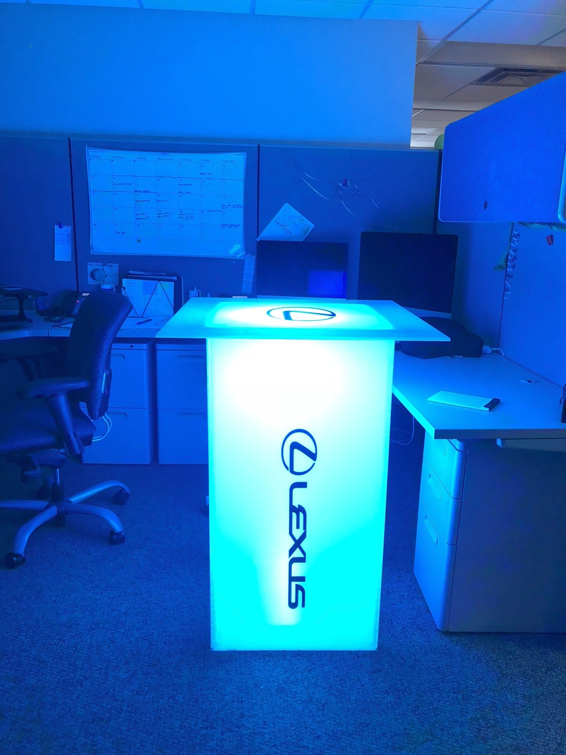 Blue-lit Lexus display pedestal in an office, with chairs and a whiteboard in the background.