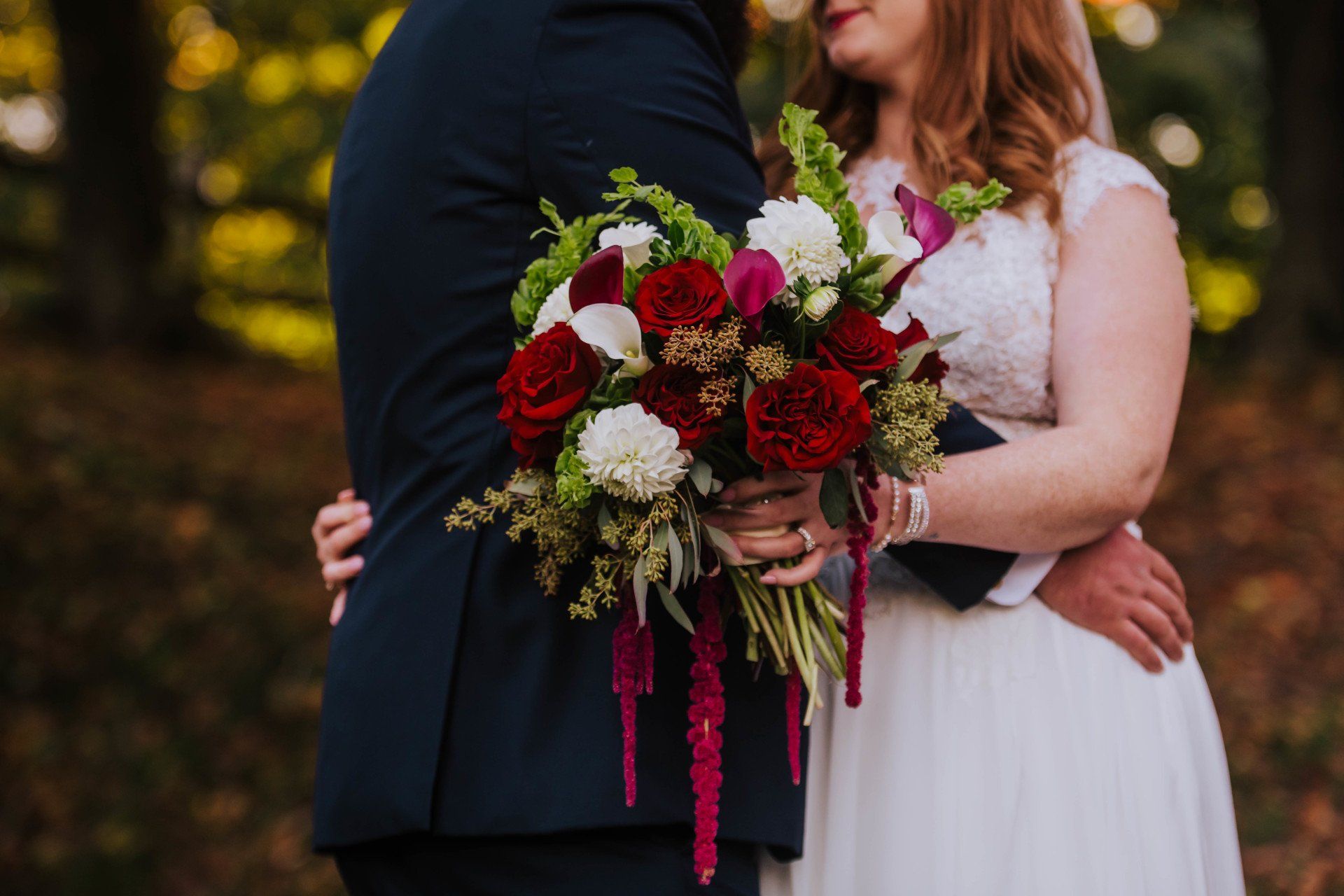 Wedding couple embracing, holding a red-and-white bouquet outdoors in autumn.