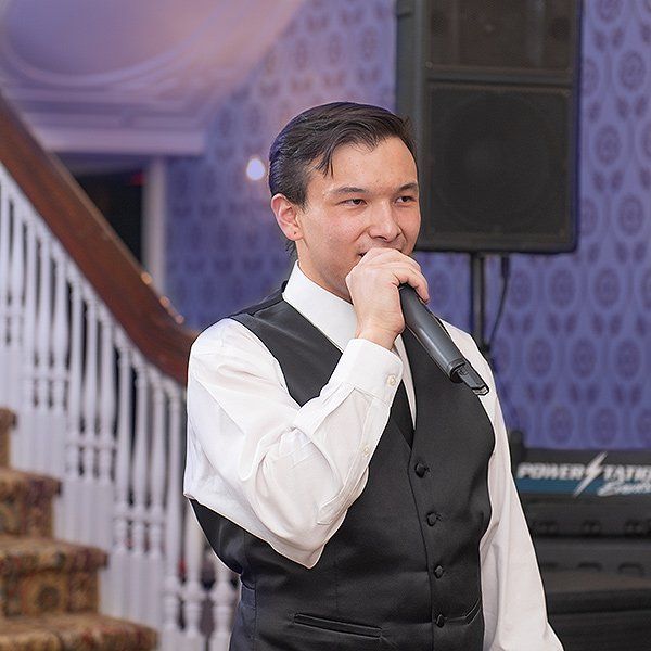Man in white shirt and black vest speaking into a microphone at an indoor event
