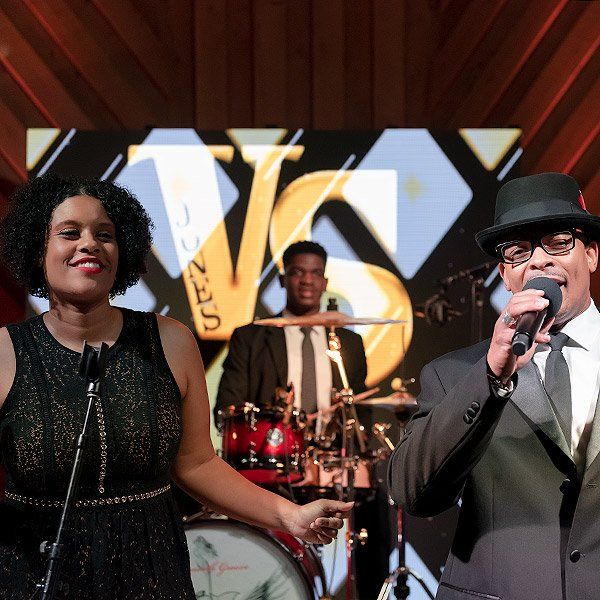 Singer and drummer performing on stage with a large gold logo backdrop