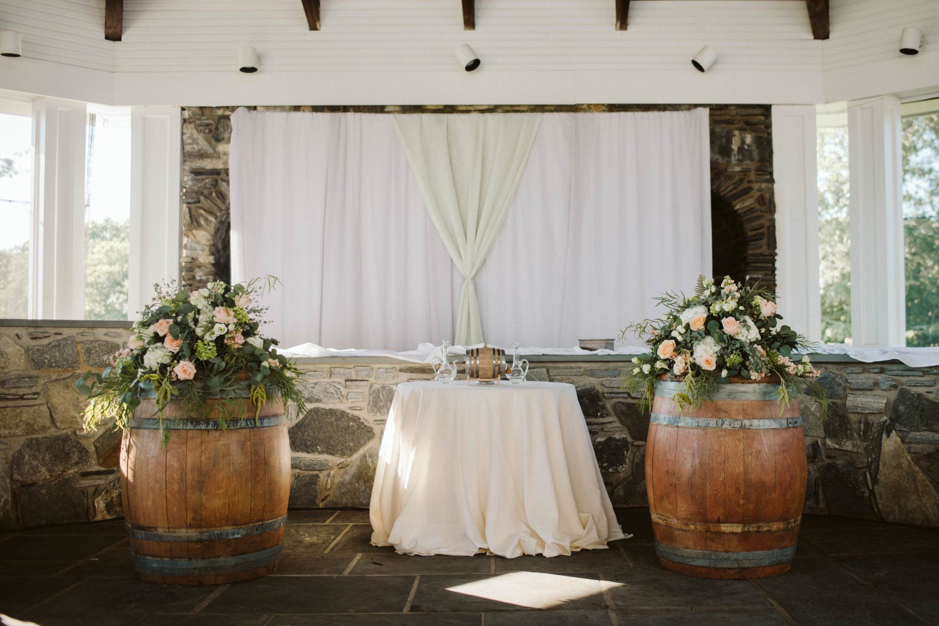 White draped wedding backdrop with small round table, floral arrangements, and two wooden barrels in a garden pavilion