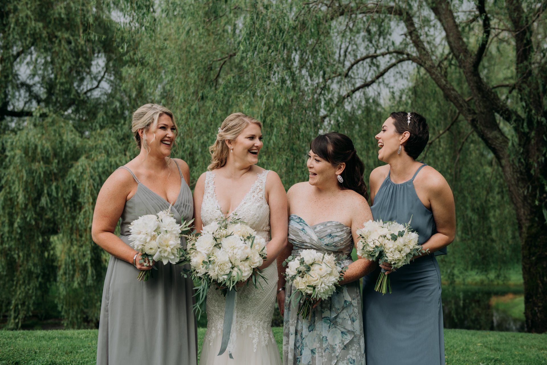 Four women in formal dresses hold white bouquets outdoors in front of leafy trees.